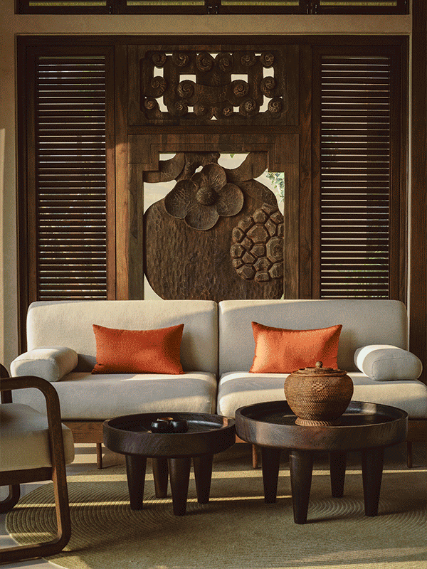 Living room with a beige sofa, two orange pillows, two round black coffee tables, a woven vase, and a wooden wall hanging with carved floral and hexagonal patterns.