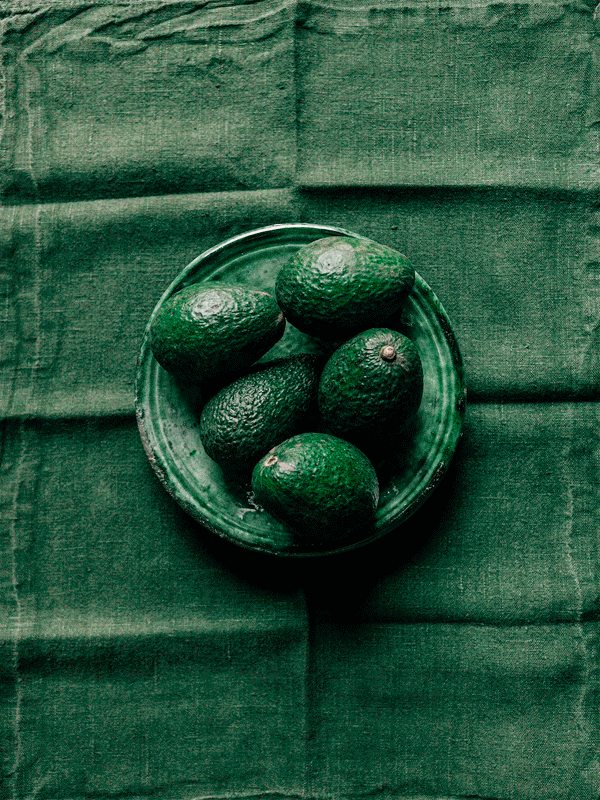 Six avocados in a glass bowl on a green fabric background.