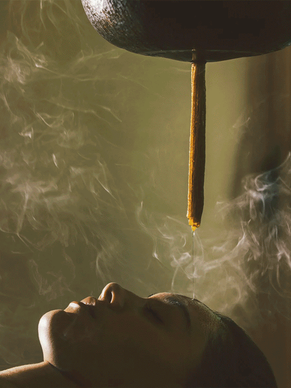 A woman receives an Ayurvedic shirodhara treatment, where warm oil is gently poured onto her forehead from a tall vessel, with steam or incense smoke surrounding her.