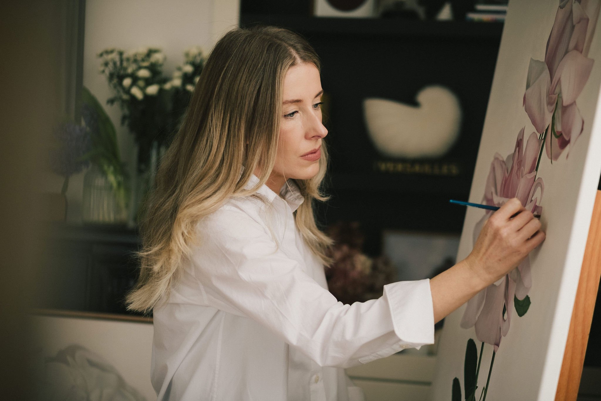 A woman with long blonde hair is painting a floral canvas. She is wearing a white shirt and is holding a paintbrush, working on a pink flower.