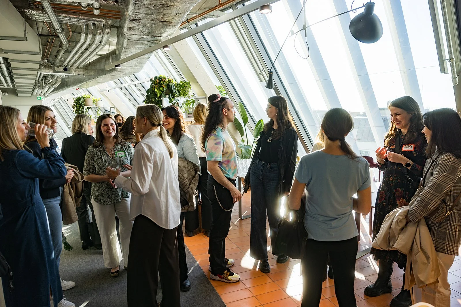 A group of women socializing and talking in a brightly lit indoor space with large windows and plants.