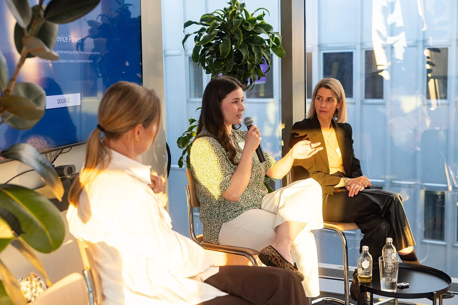 Three women seated in a panel discussion, one speaking into a microphone, in a modern room with large windows and indoor plants.