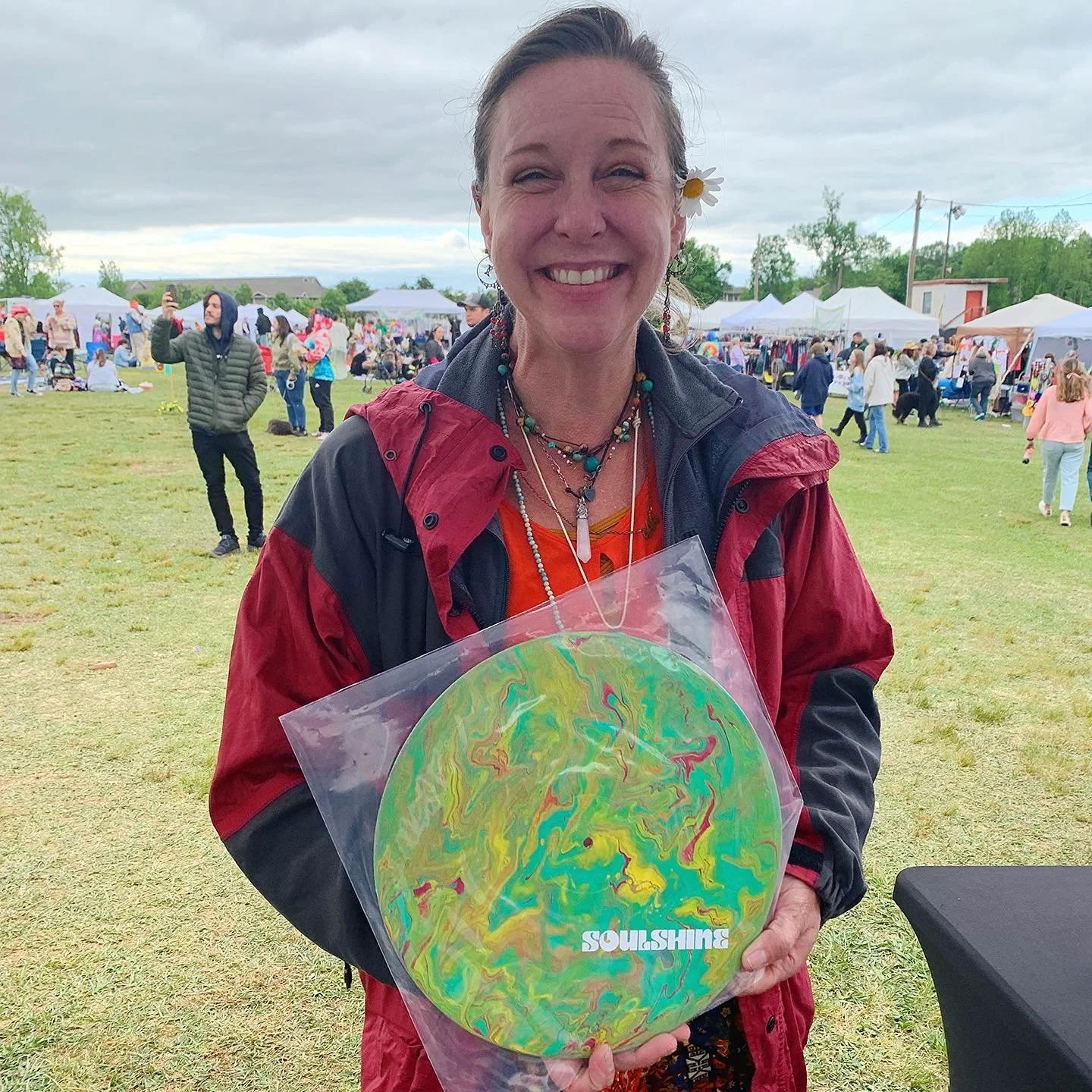 A smiling woman wearing colorful jewelry and a flower in her hair, holding a vinyl record with the word "SOULSHINE" on it, at an outdoor event with tents and people in the background.