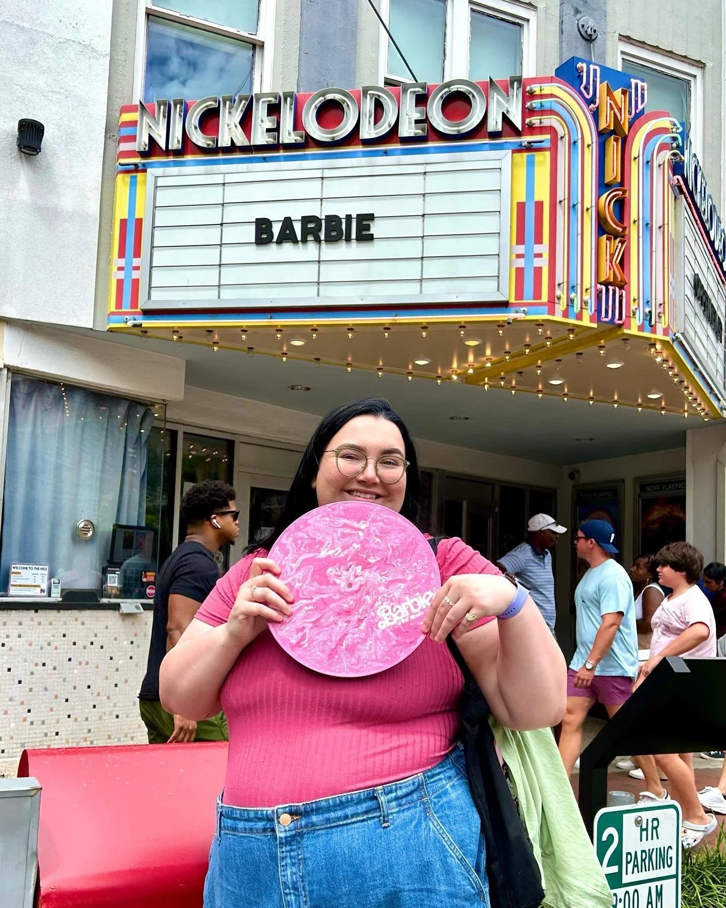 A woman smiling and holding a pink bubblegum ice cream outside of a Nickelodeon theater. The theater marquee displays the name 'Barbie'.
