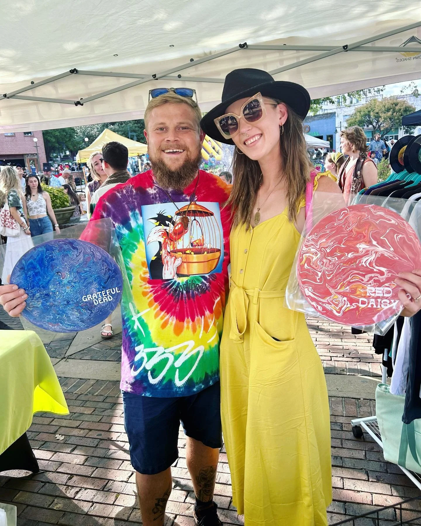 Two smiling people holding vinyl records at an outdoor event, with tents and other attendees in the background.