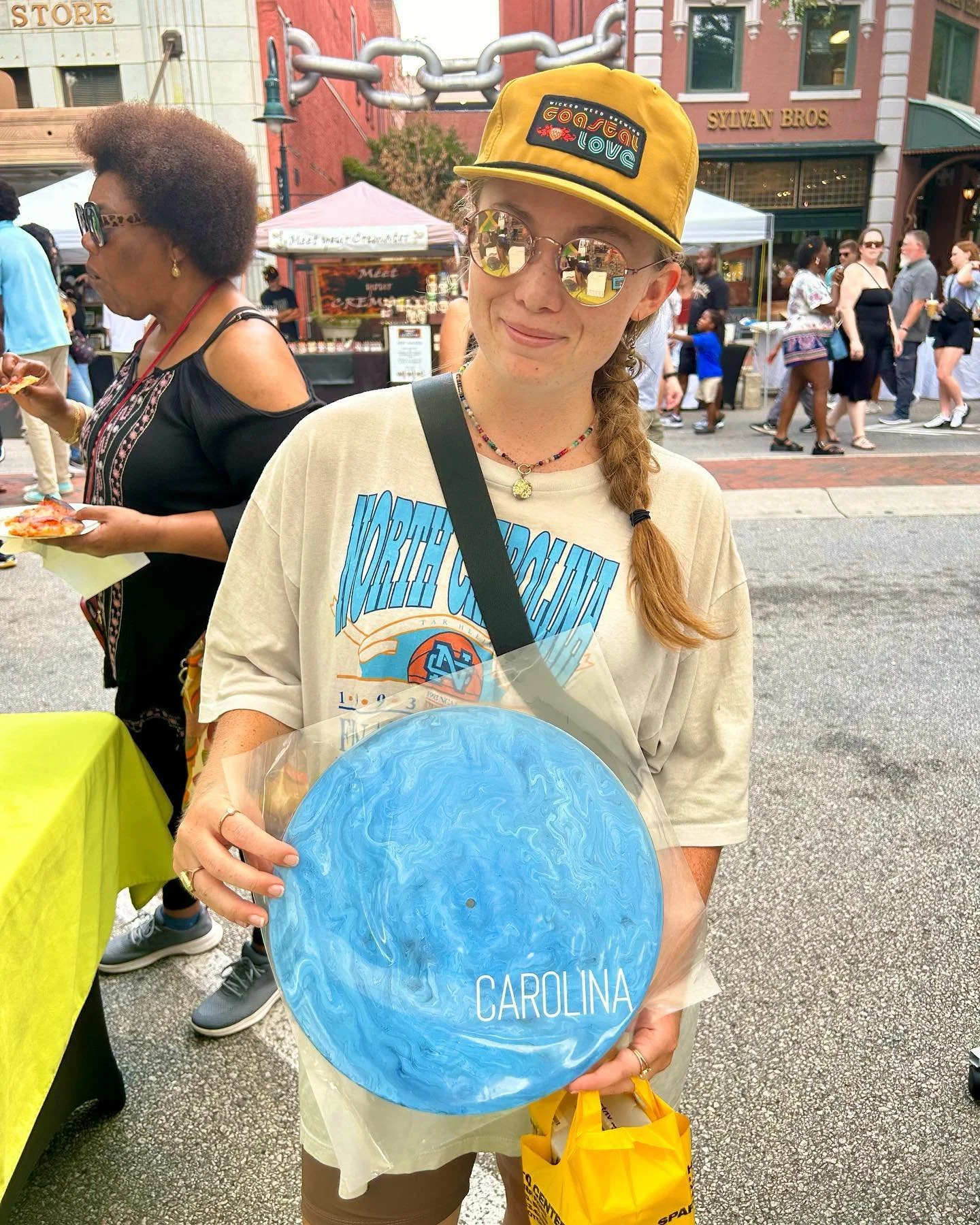 A young woman wearing sunglasses, a yellow hat, and a North Carolina T-shirt is holding a blue marbled disc labeled "CAROLINA" at an outdoor event with many people, tents, and a brick building in the background.