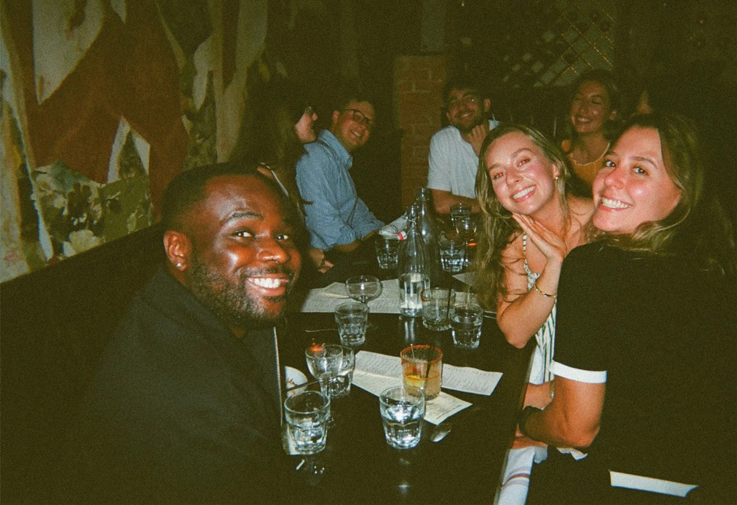Group of six people smiling and enjoying drinks at a Bar Sotto meal, seated around a table with glasses and bottles.