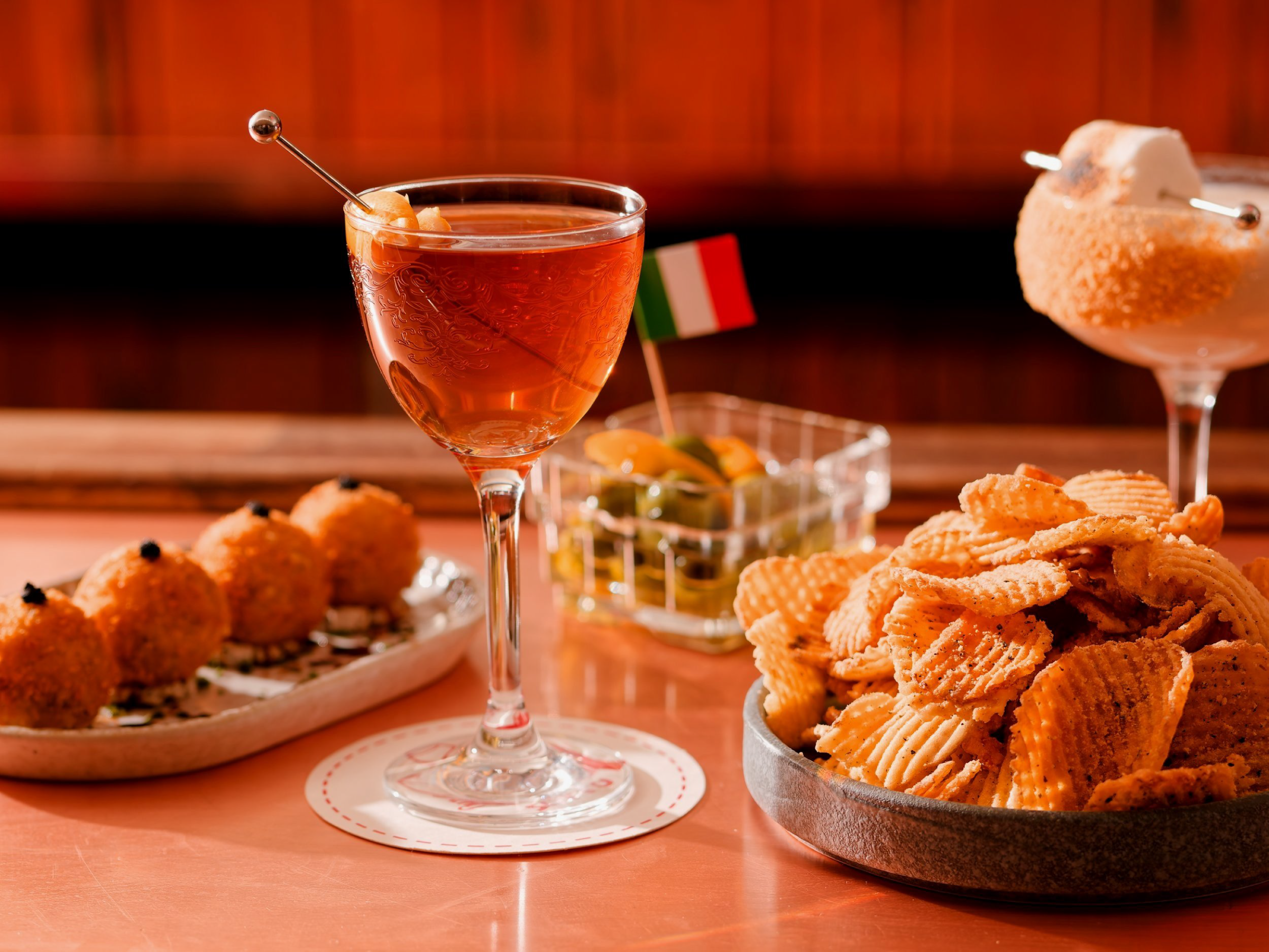 Various Bar Sotto snacks and drinks, including a glass of red wine or cocktail with an olive, a bowl of potato chips, a shot of a layered cocktail garnished with a cookie, a bowl of fried croquettes, and a small Italian flag in the background.