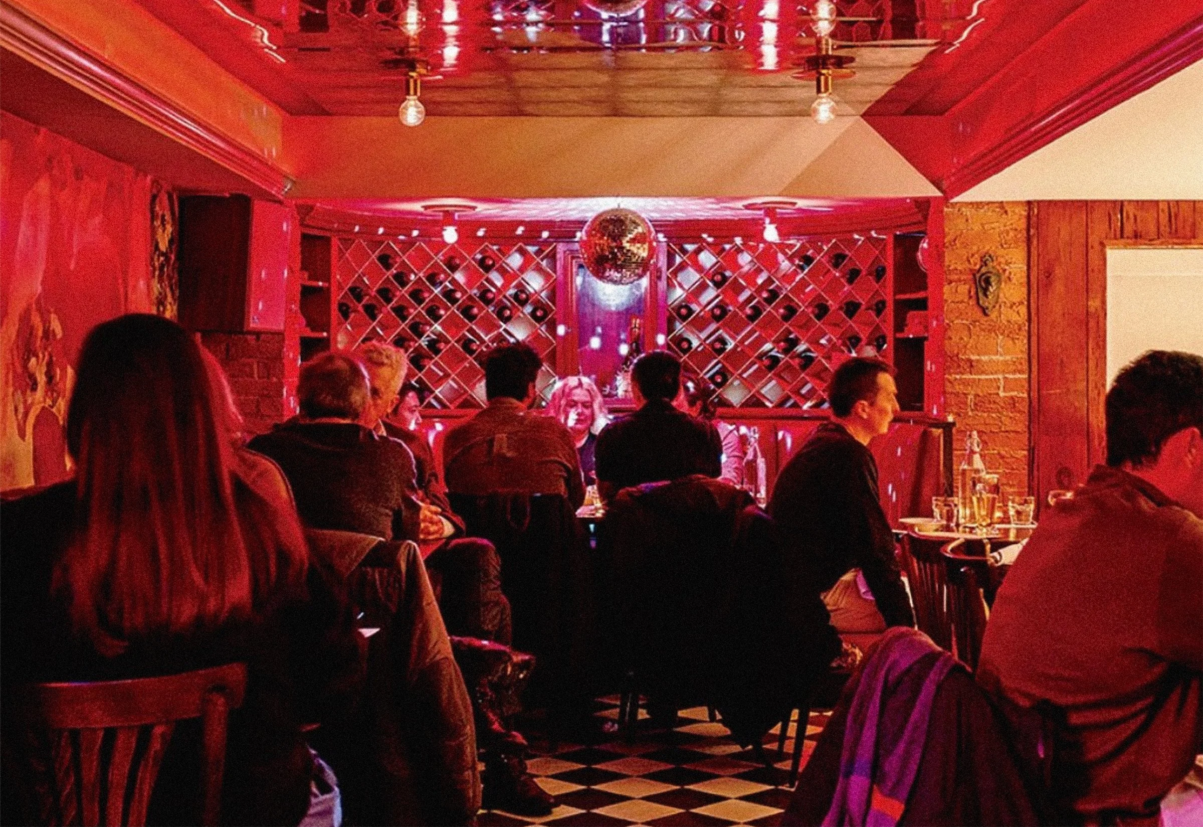 People sitting at Bar Sotto tables in a dimly lit bar with red lighting, wine bottles on shelves, and a disco ball hanging from the ceiling.
