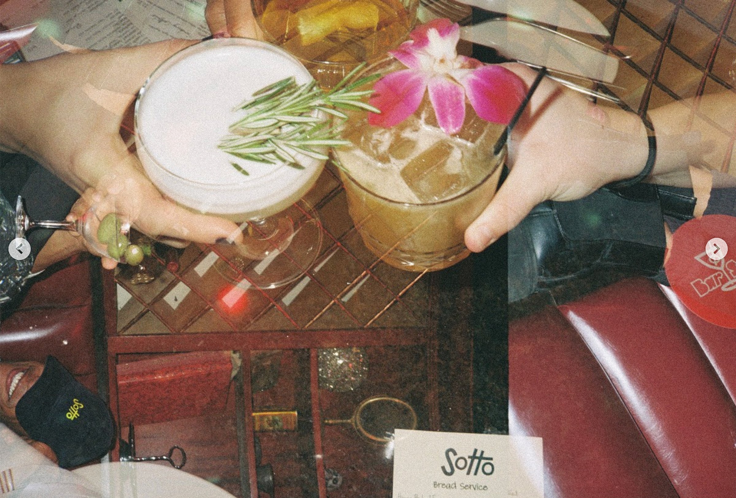 Two hands holding cocktails, one with a frothy drink garnished with rosemary and the other with a drink garnished with pink and white flowers, over a bar table.