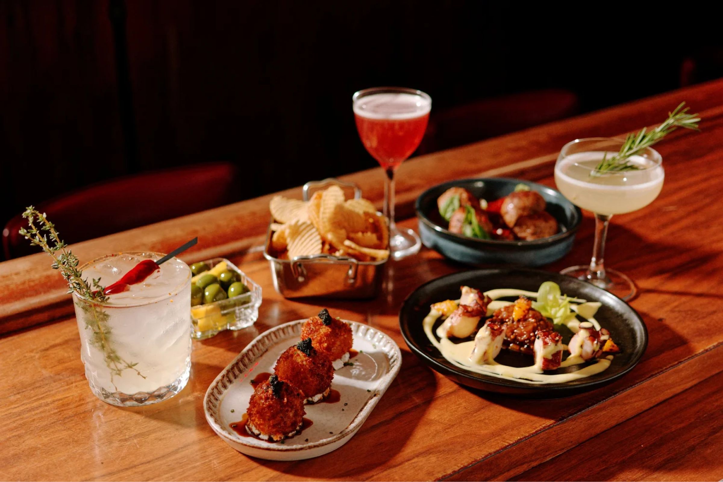 Assorted appetizers and drinks on a wooden table, including a cocktail with a sprig of rosemary, a small dish of olives, a bowl of potato chips, a plate of fried croquettes, a plate of sushi rolls with sauce, and two cocktail glasses with garnishes.
