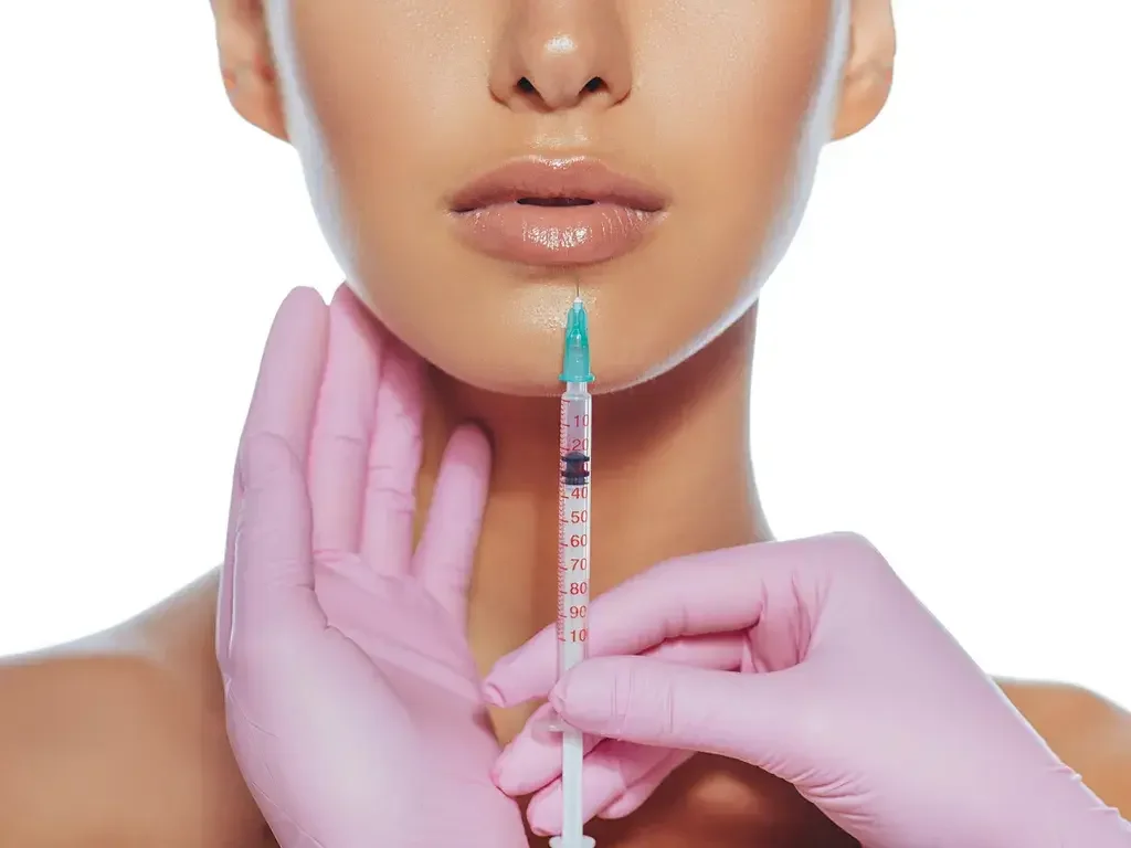 Close-up of a woman receiving a cosmetic injection under her chin, held by hands wearing pink gloves on a white background.