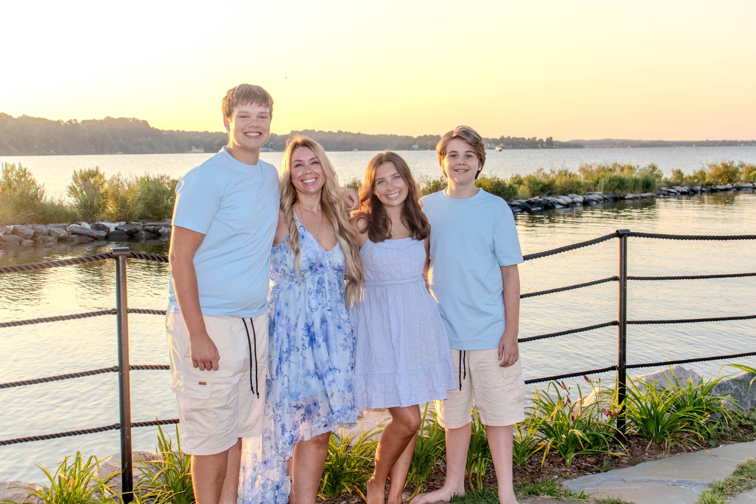 A family of four with three children and one adult mother, standing on a lakeside promenade at sunset, smiling and posing for a photo.
