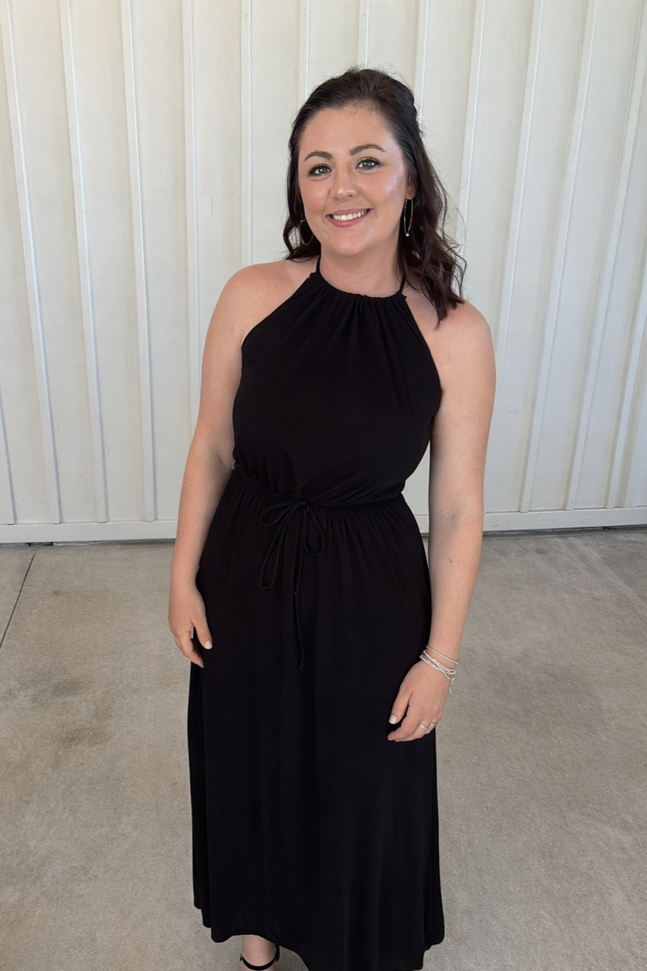A woman with dark brown hair and hoop earrings wearing a black sleeveless dress, standing against a light-colored wooden wall, smiling at the camera.