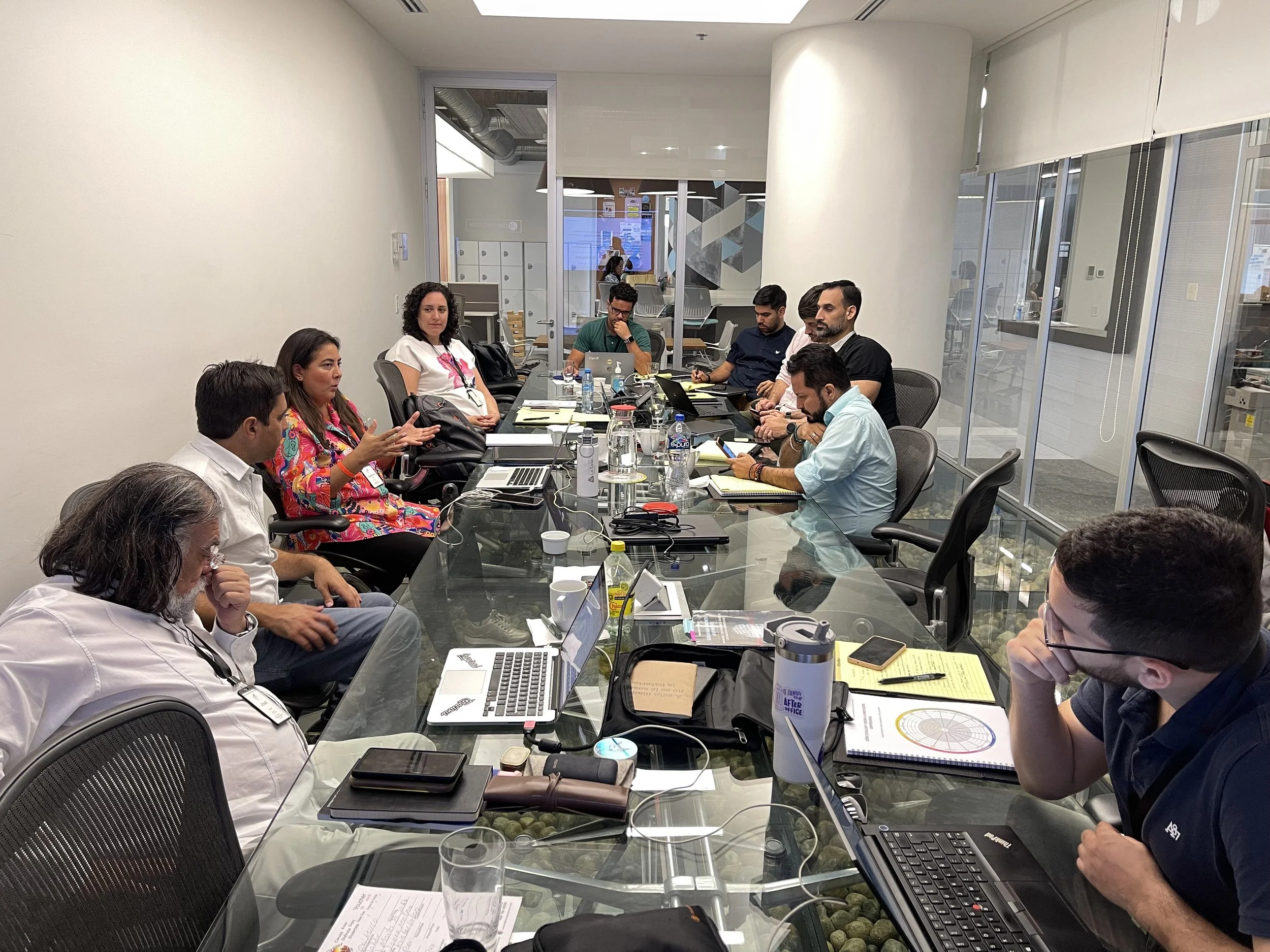 Reunión de trabajo en una sala de conferencias con varias personas sentadas a la mesa, algunos con computadoras portátiles y documentos, discutiendo y tomando notas.