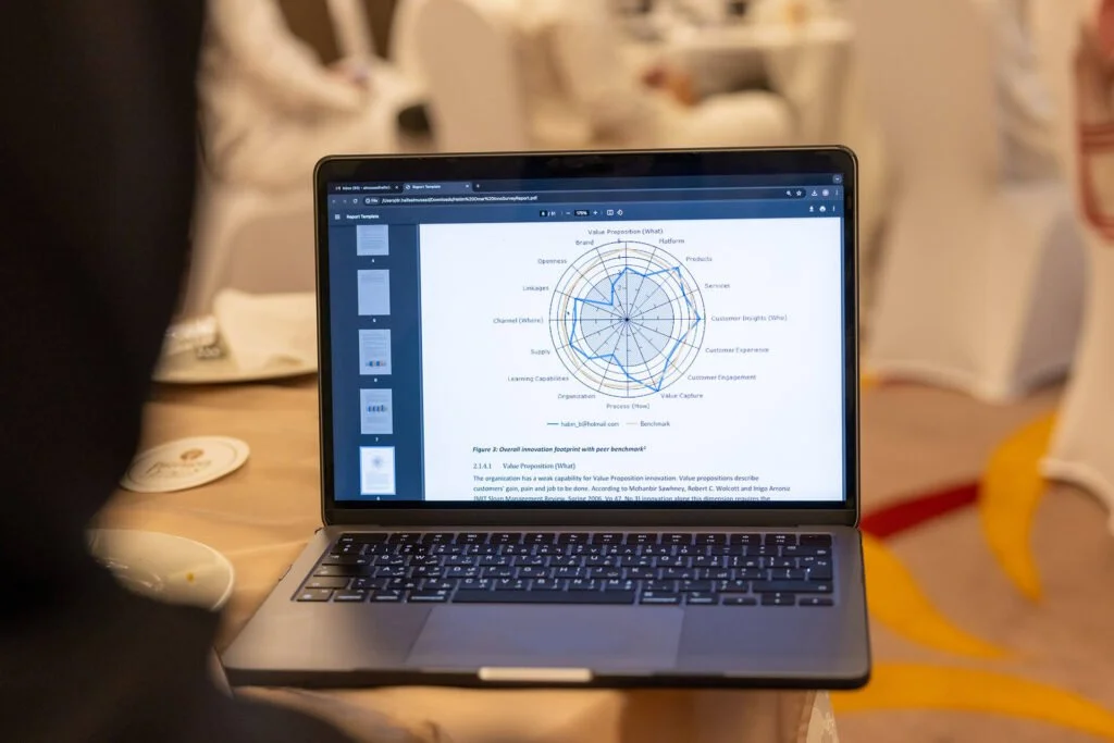 Laptop en una mesa mostrando un gráfico circular en una presentación o reporte, con otras personas en el fondo en un ambiente de reunión o conferencia.