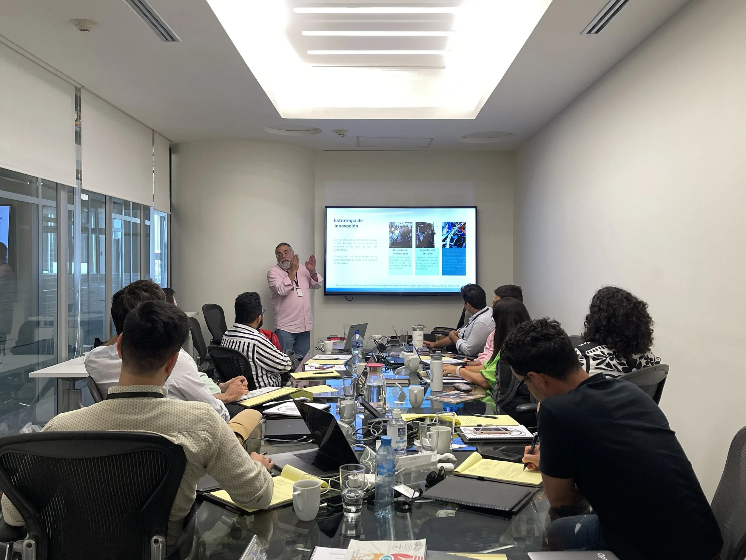 Reunión en una sala de conferencias con un presentador explicando una diapositiva a un grupo de personas sentadas en una mesa larga, con laptops, cuadernos y bebidas.