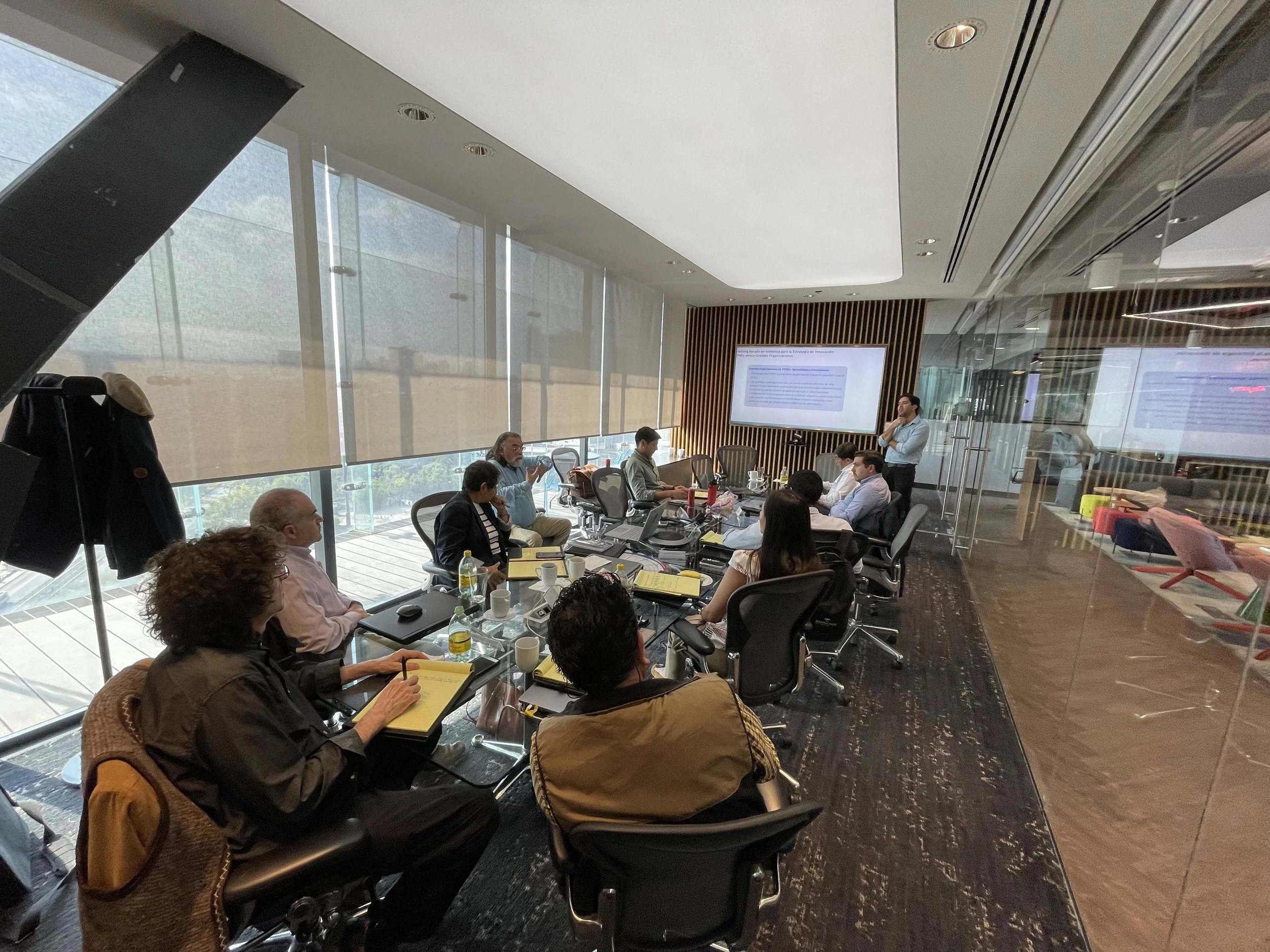 Reunión de oficina con varias personas sentadas alrededor de una mesa de conferencias, una persona de pie frente a una pantalla de presentación.