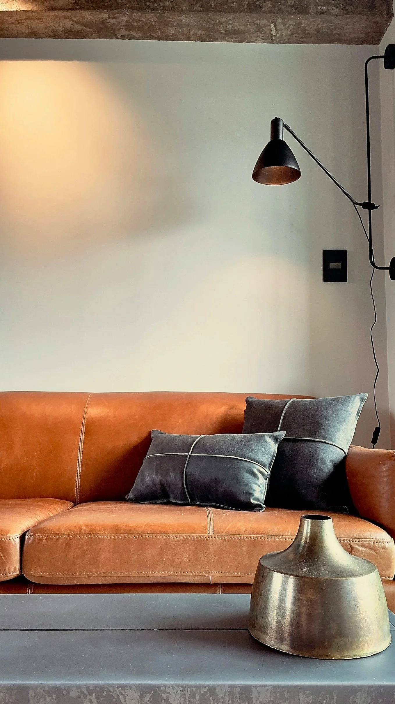 Living room with a brown leather sofa, black pillows, a gray coffee table with a metallic vase, a black wall light with an adjustable arm, and a white wall with a black switch.