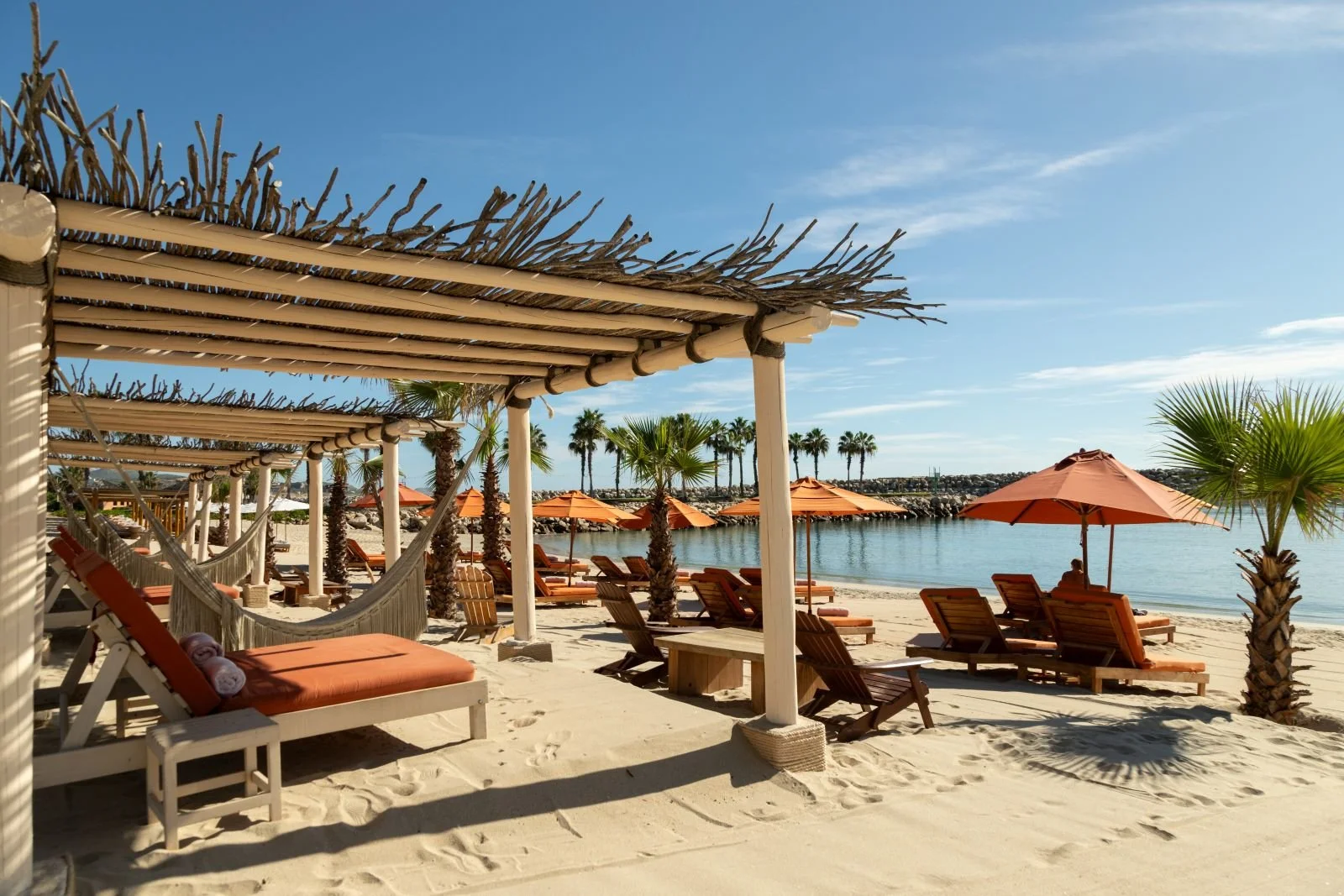 Beachside lounge chairs with orange umbrellas and wooden cabanas along a sandy shoreline, overlooking calm water and palm trees against a blue sky.