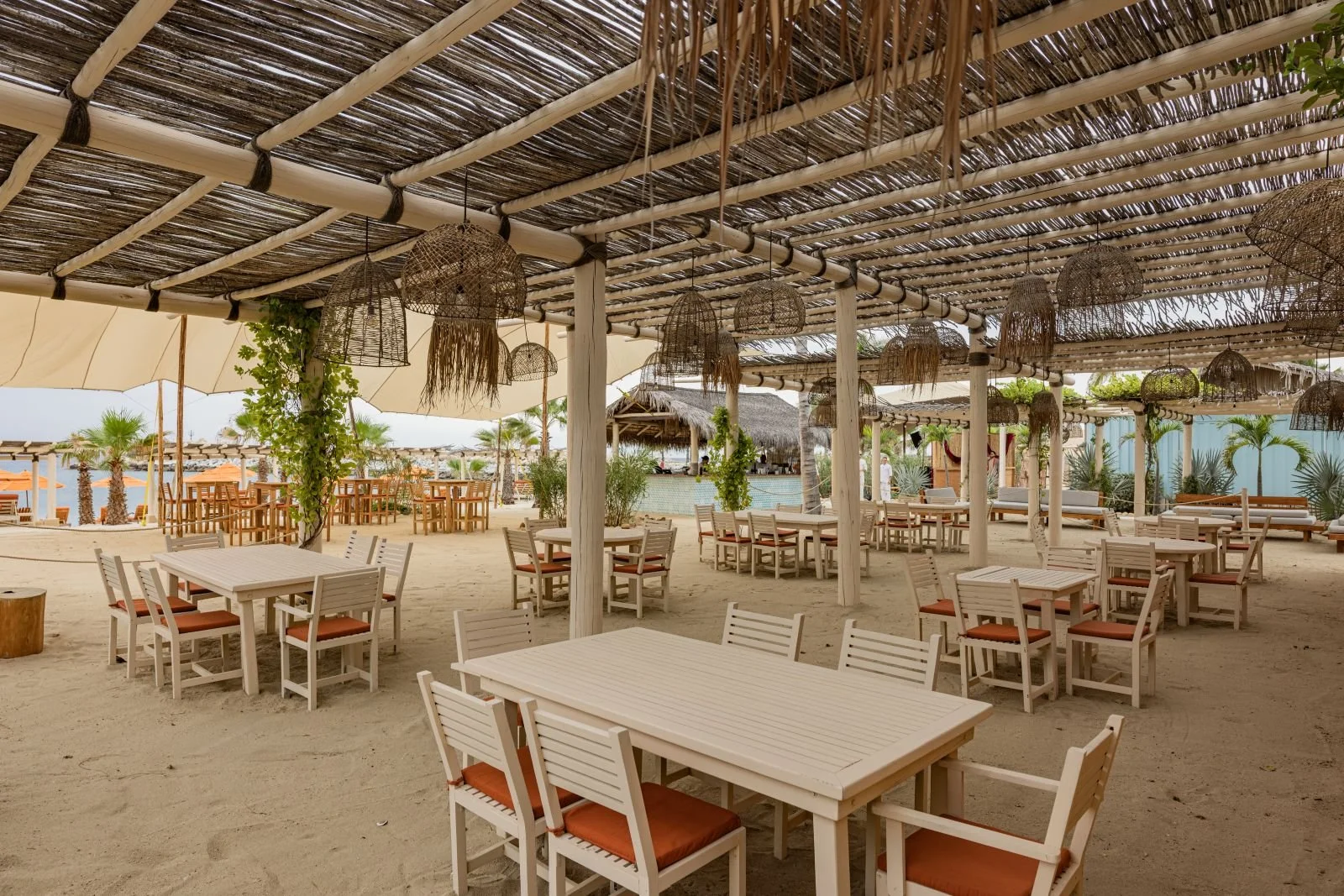 Empty outdoor beach restaurant with white wooden tables and chairs on sandy ground, surrounded by palm trees, with woven pendant lights hanging from a thatched roof structure.
