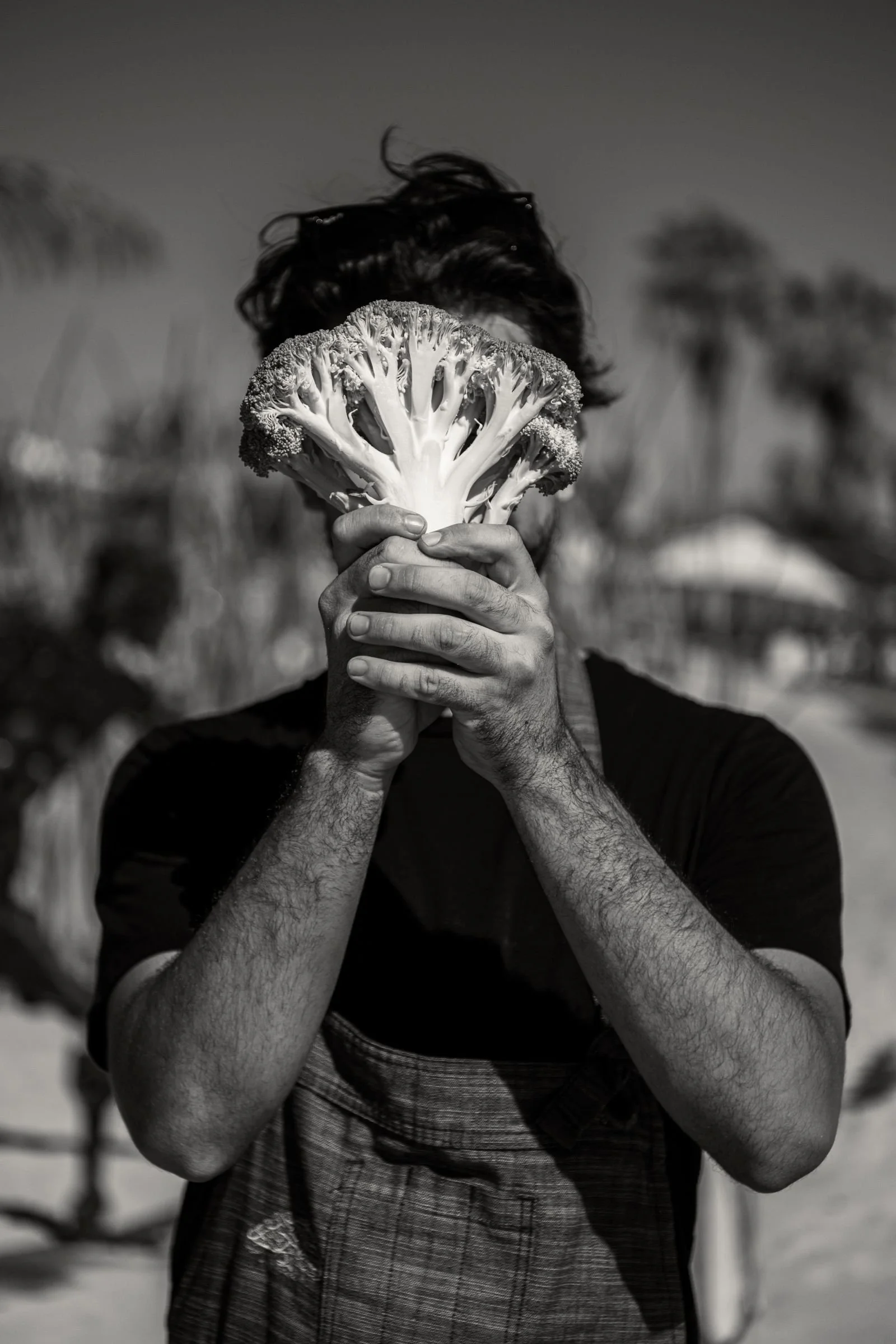 Angel Camarena, Velero's chef, holding a broccoli in front of their face, obscuring it, outdoors on a sunny day.