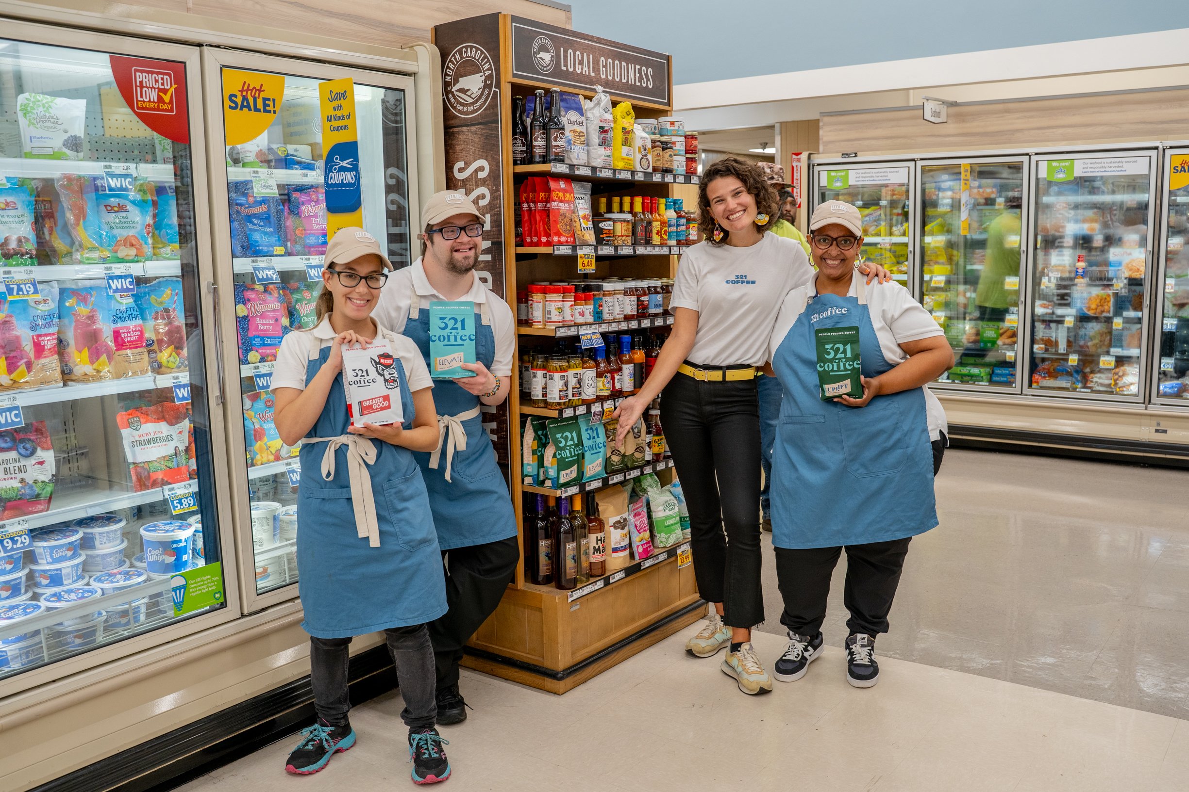 Lindsay stands with 3 baristas in blue aprons in the aisle of a Food Lion with 321 Coffee bags on the shelf.