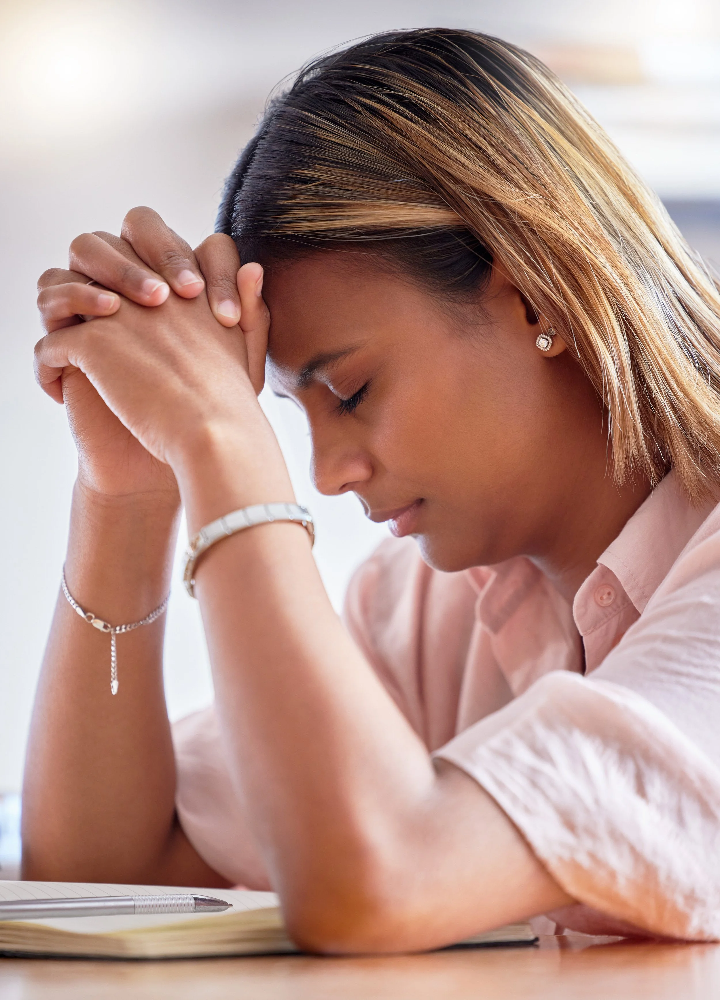 A woman with light brown and blonde hair, eyes closed, sitting at a desk with her hands clasped on her forehead, a notebook and pen in front of her, appearing stressed or tired.