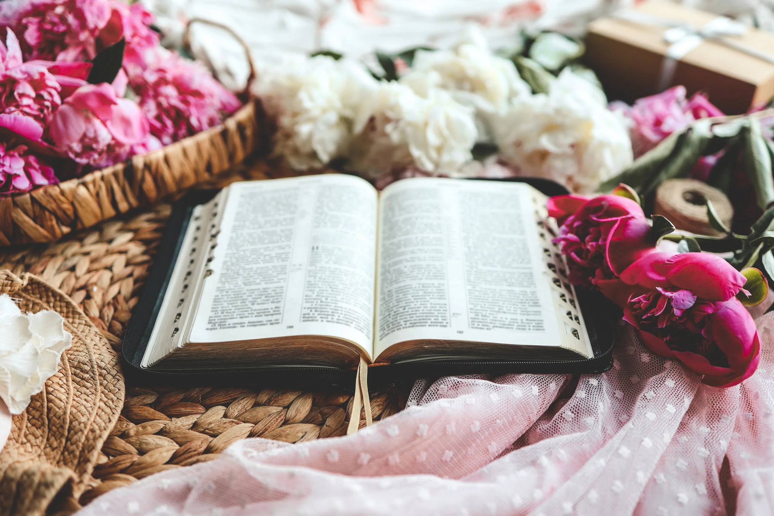 Open Bible surrounded by pink and white flowers, woven baskets, and pink fabric.