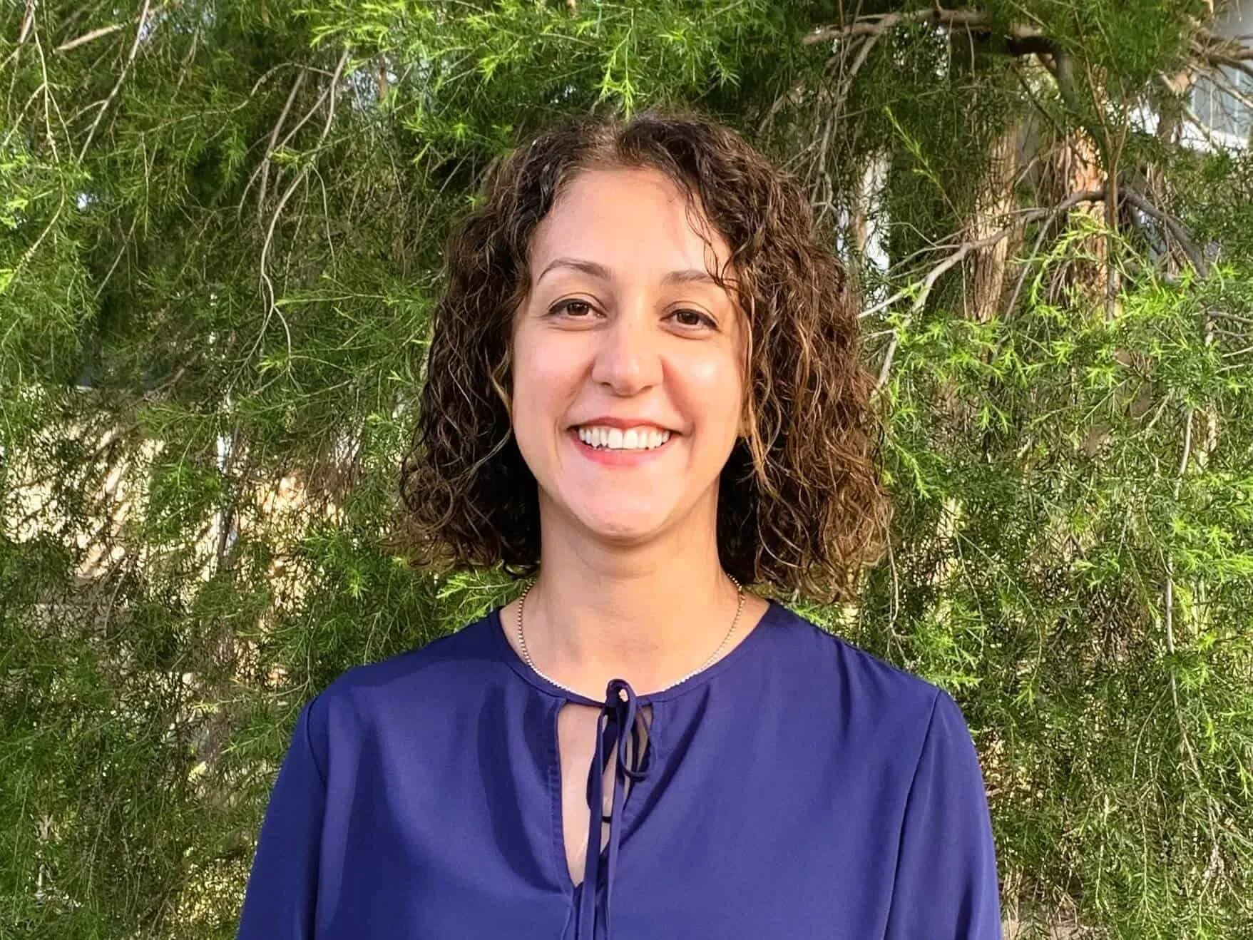 Mona Ghafourian in a blue shirt smiling outdoors in front of green leafy trees in Bella Vista.