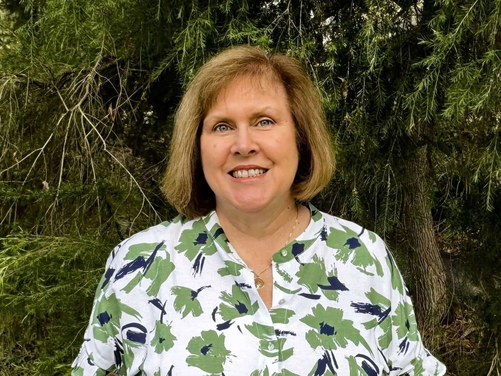 Lynne-Maree Crain in a green floral top smiling outdoors in front of green leafy trees in Bella Vista.