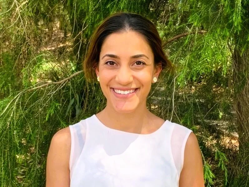 Monica Asimus in a sleeveless white top smiling outdoors in front of green leafy trees in Bella Vista.