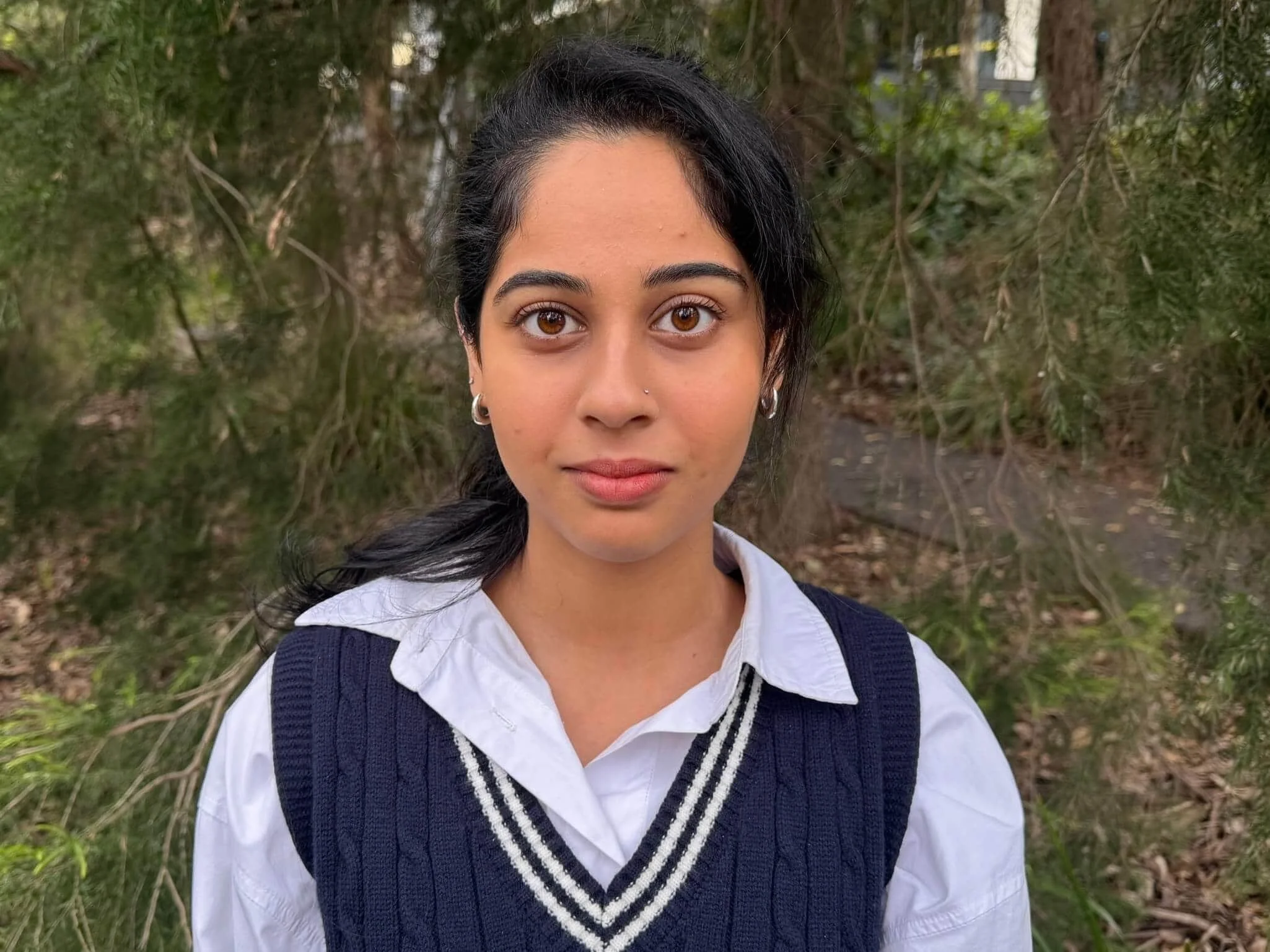 Maha Baig in a white shirt and navy vest, smiling outdoors in front of green leafy trees in Bella Vista.