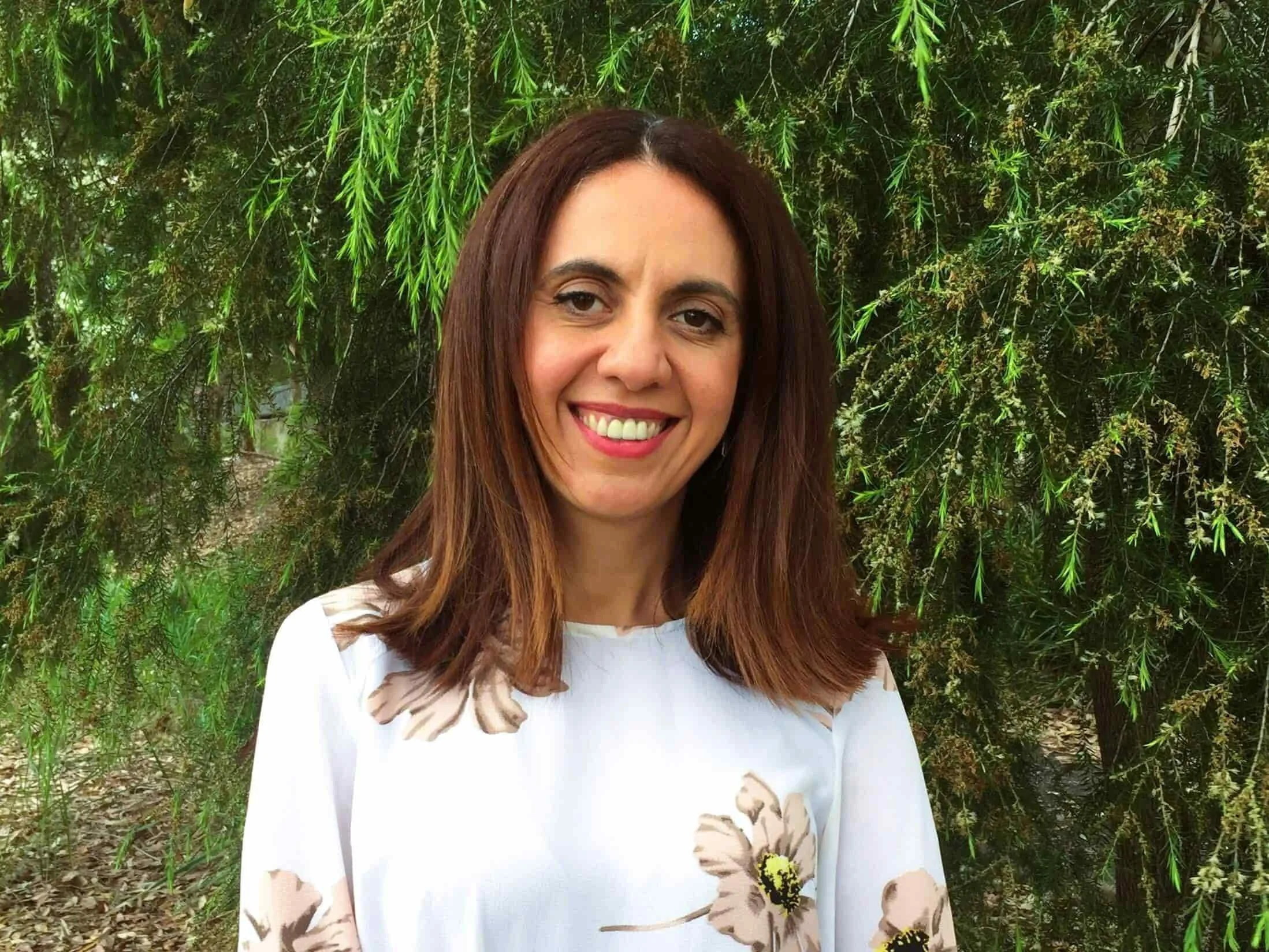 Natalie Foti in a pink and white floral shirt smiling outdoors in front of green leafy trees in Bella Vista.