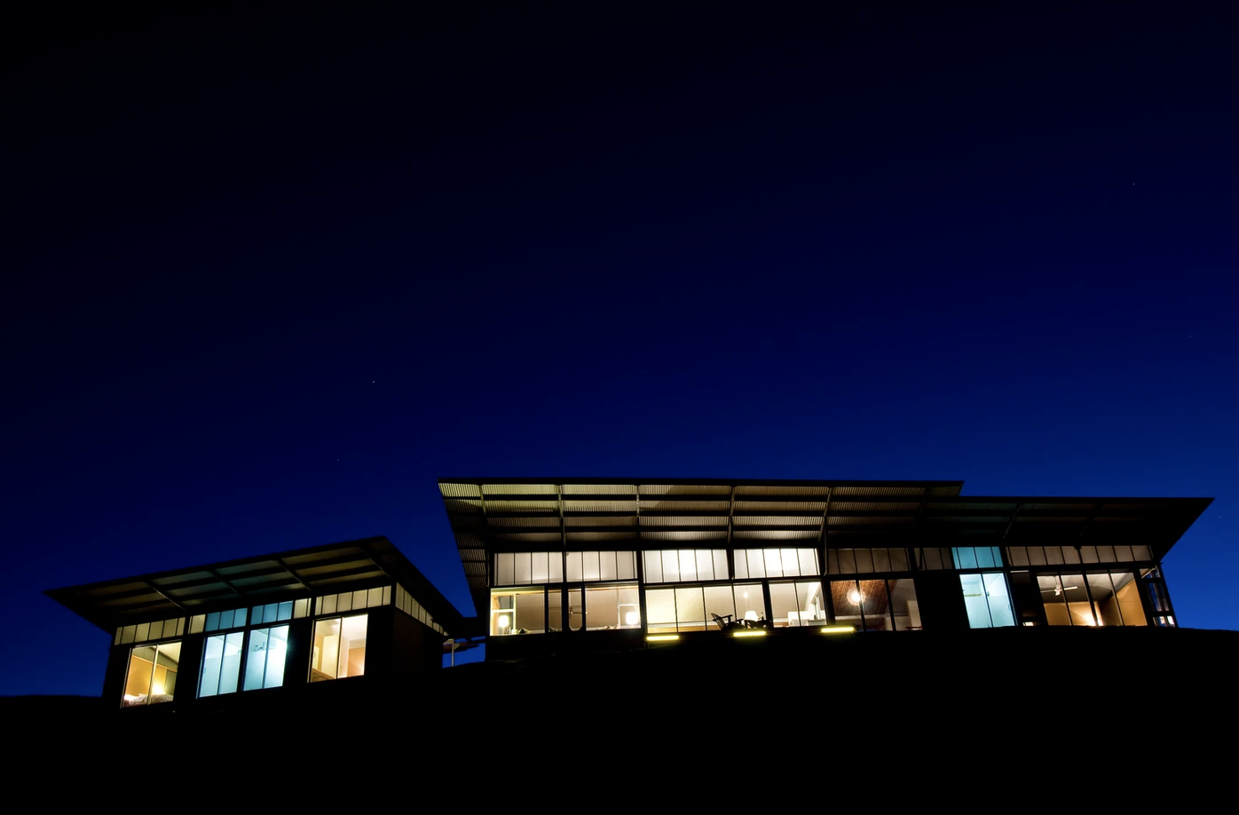 Modern house with large windows illuminated at night, overlooking a dark sky.