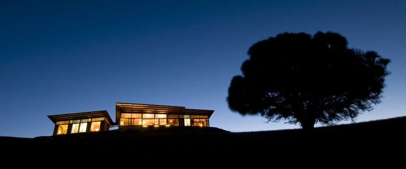 A modern house with illuminated windows on a hill at dusk, with a large tree nearby and a darkening sky in the background.