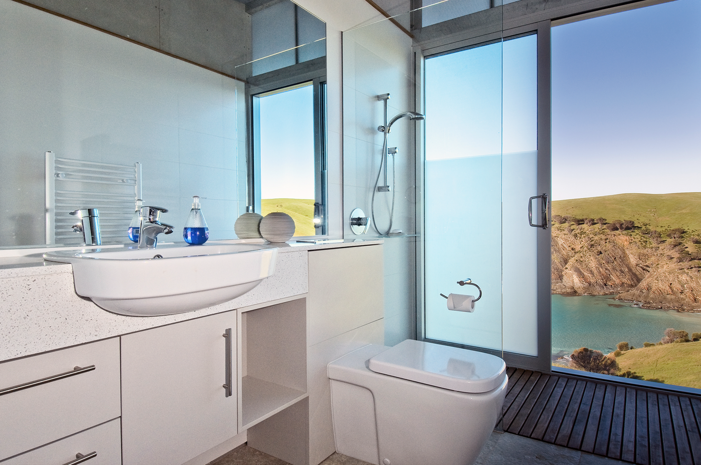 Modern bathroom with large window showing scenic ocean view, featuring a white sink, blue bottles, a toilet, and a shower with a glass door.