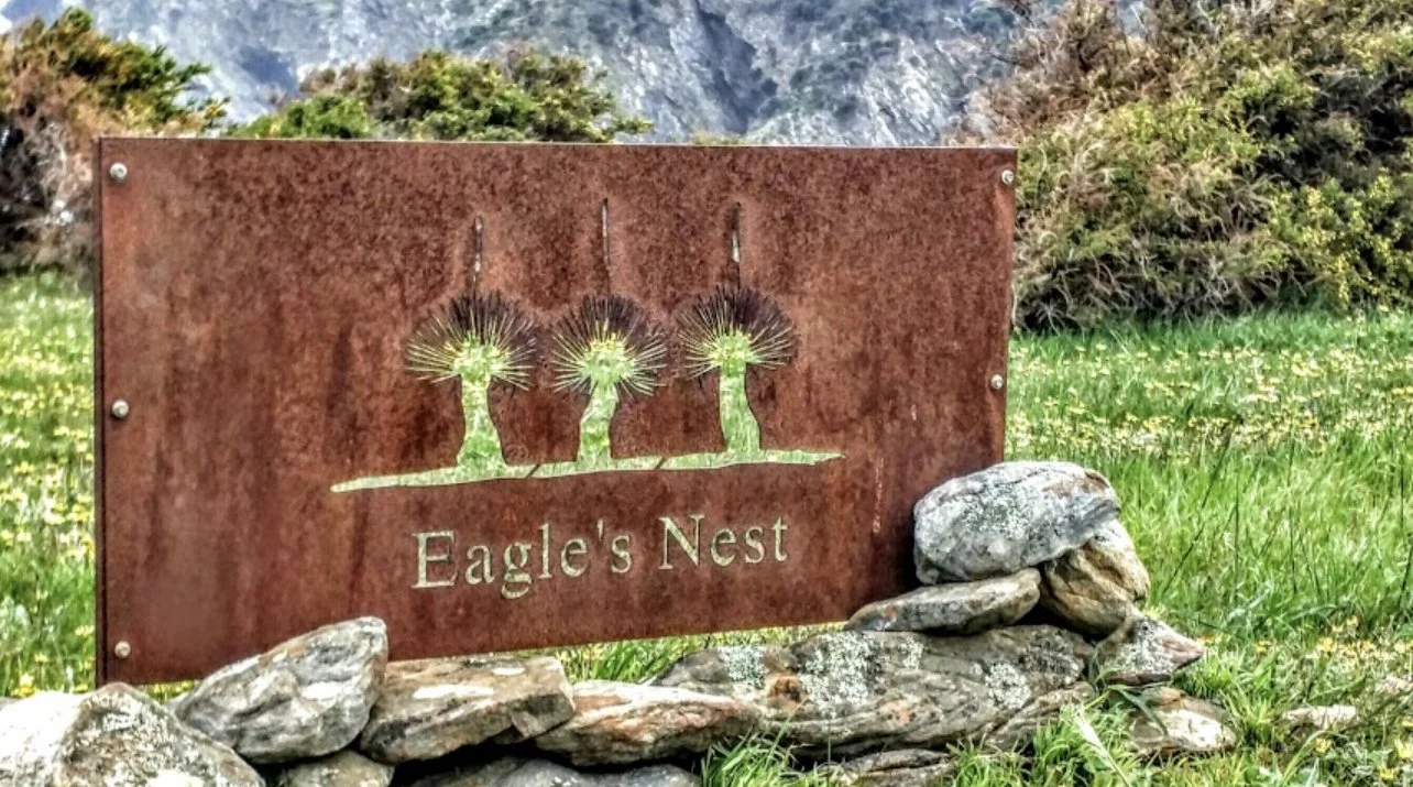 A rust-colored metal sign that says 'Eagle's Nest' with an illustration of three trees, surrounded by rocks and green grass, with mountains and bushes in the background.