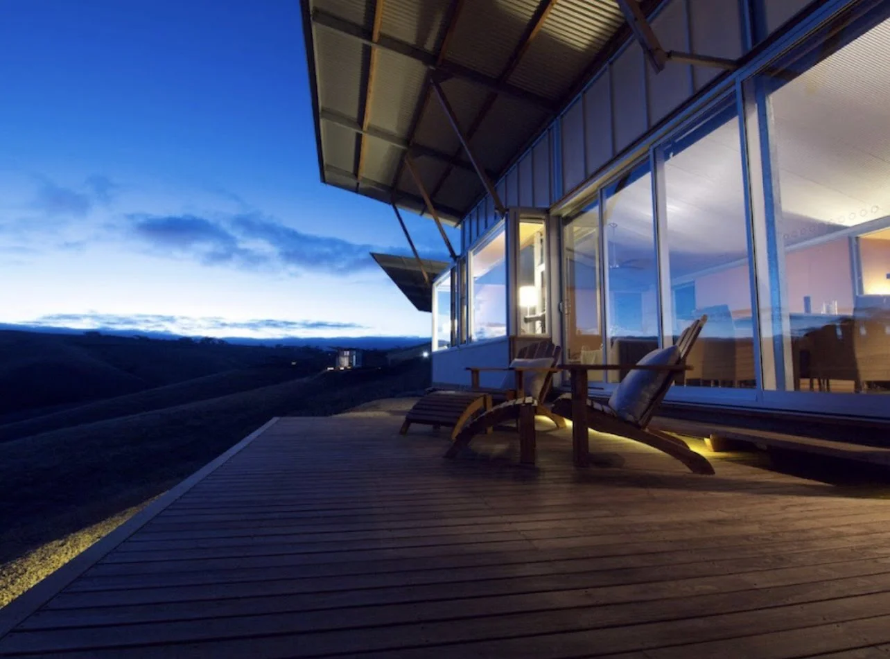 A modern house with large glass windows and a deck with two wooden lounge chairs, overlooking a dark landscape and a blue sky during twilight hours.