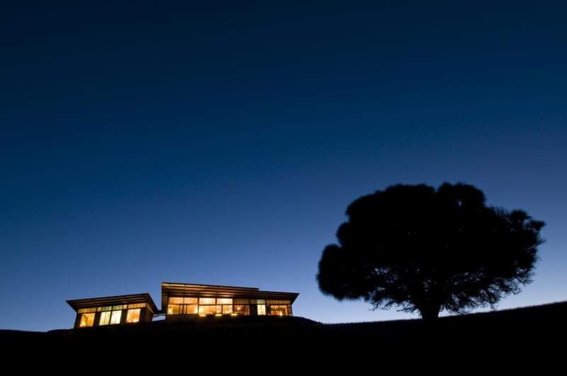 A house with illuminated windows on a hilltop next to a large tree, under a dark twilight sky.