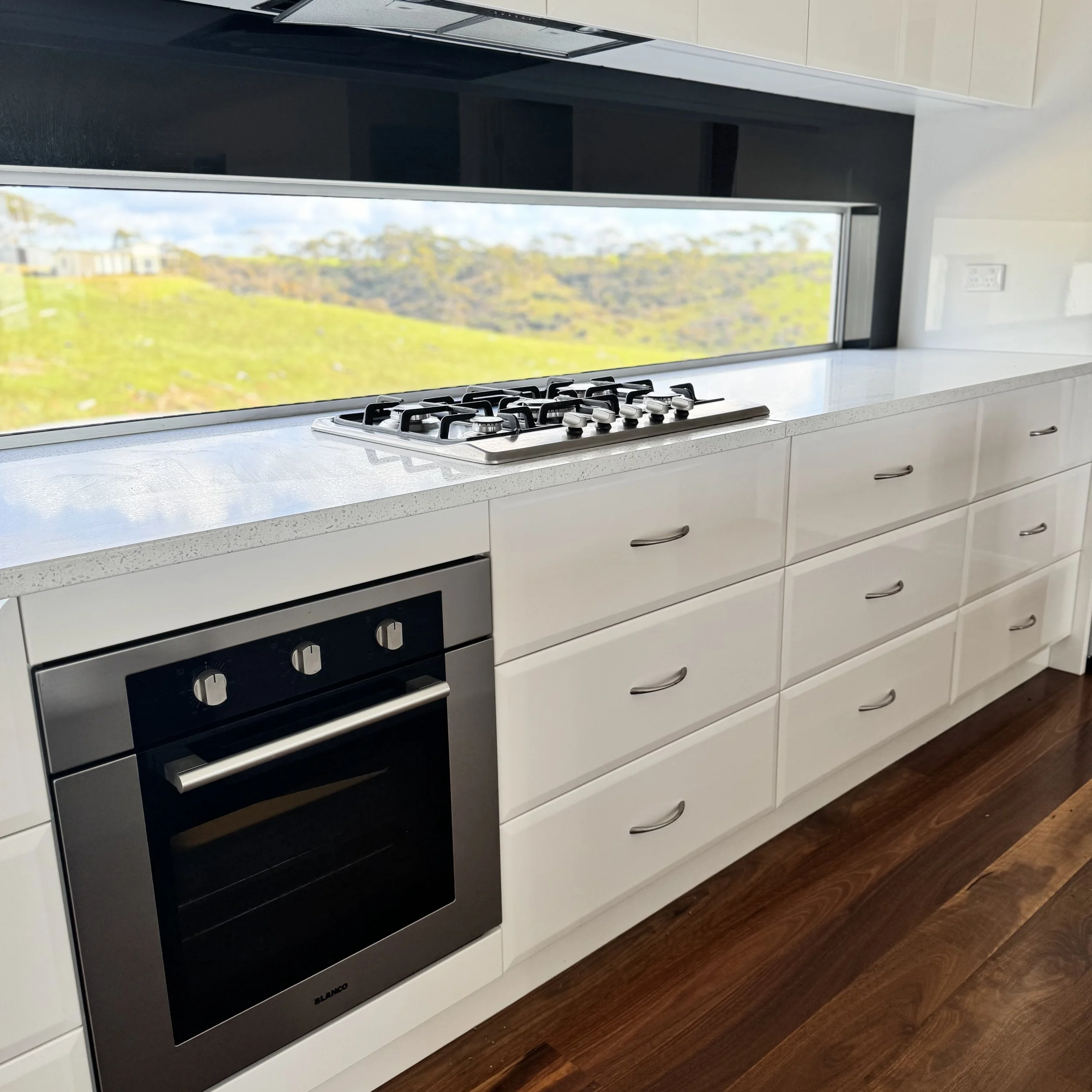 Modern kitchen with white cabinets, a stove on white countertop, a stainless steel oven, and a window showing a view of green hills.