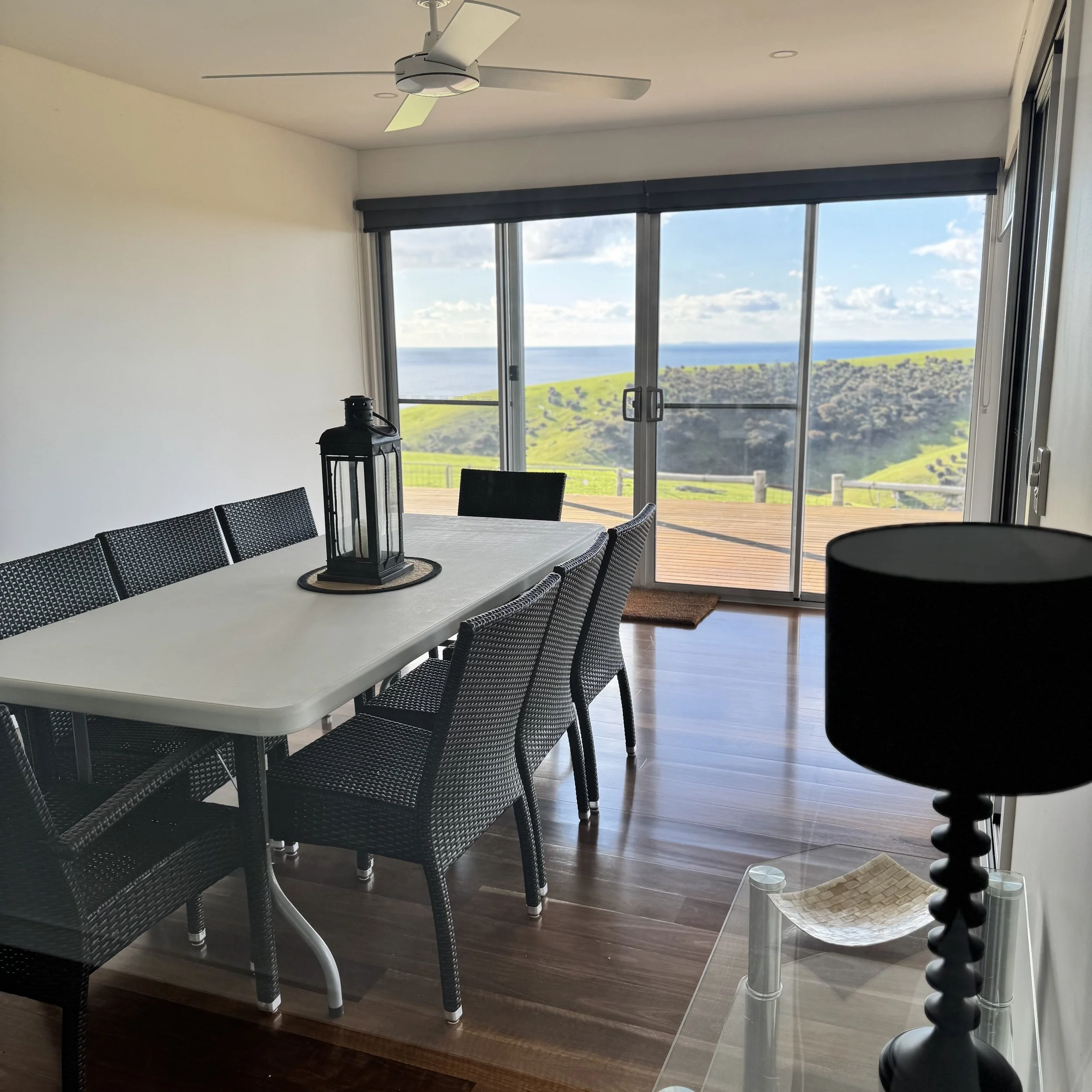 Dining room with a white table, black woven chairs, black lantern centerpiece, sliding glass doors leading to a balcony with scenic ocean and green hillside view, ceiling fan, and a black table lamp.