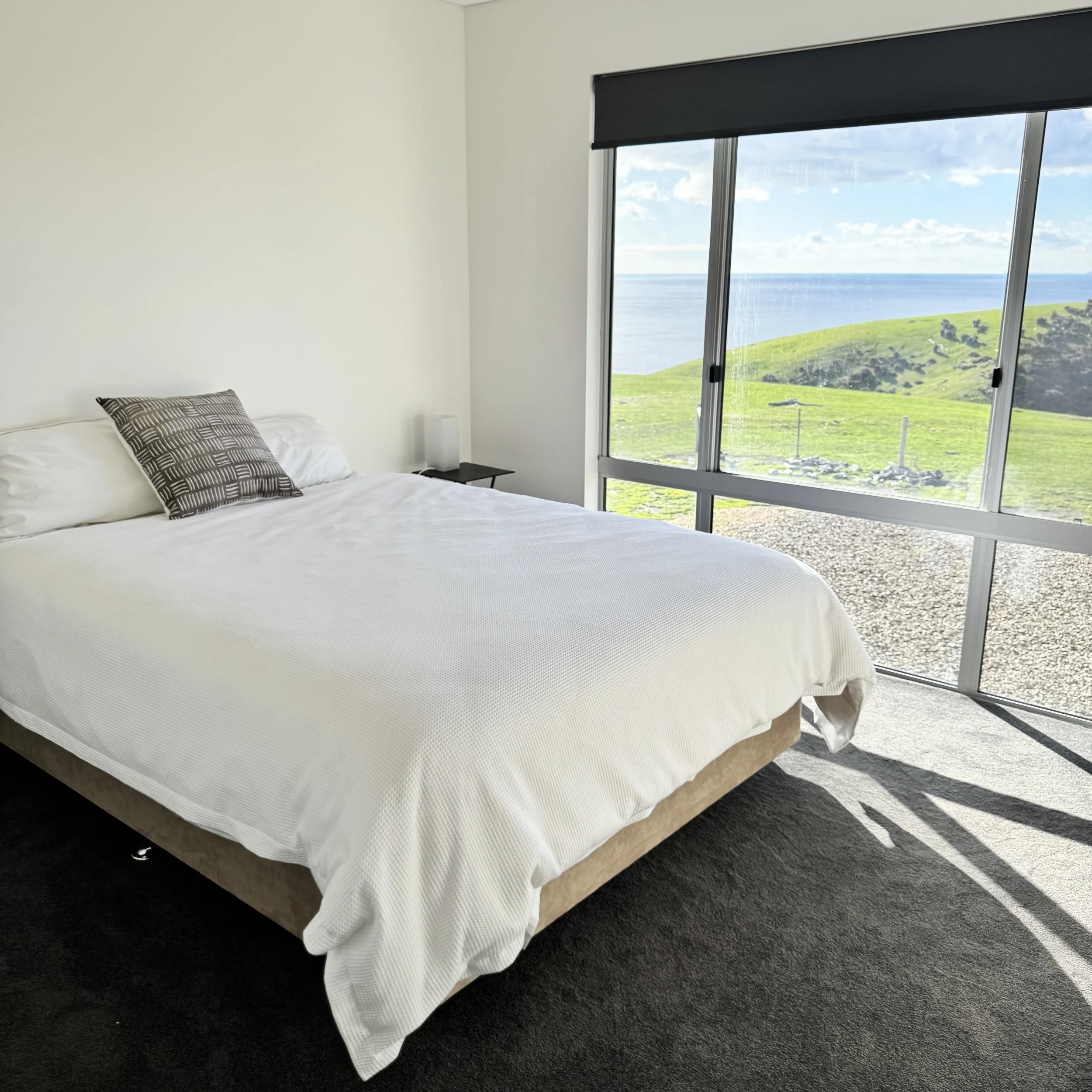 A bedroom with a large bed, white bedding, and a black and white patterned pillow, next to a window with a view of green hills and the ocean.