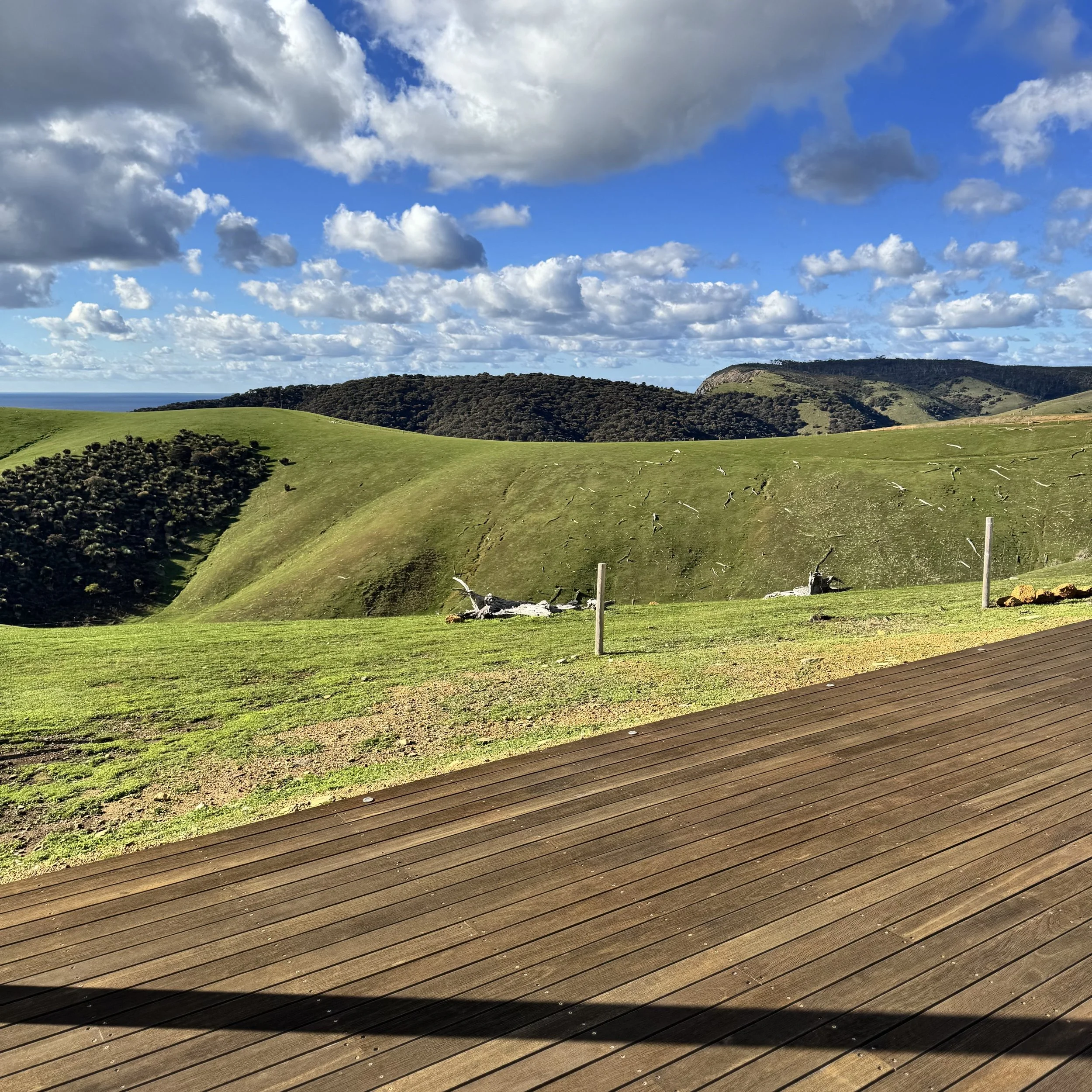 A scenic view of green rolling hills and distant mountains under a partly cloudy sky, with a wooden deck at the foreground.