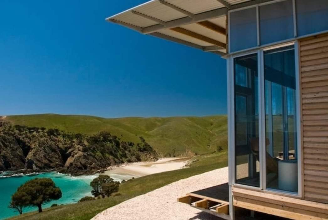 Part of a modern house with large glass windows, overlooking green rolling hills, a beach, and a turquoise ocean beyond.