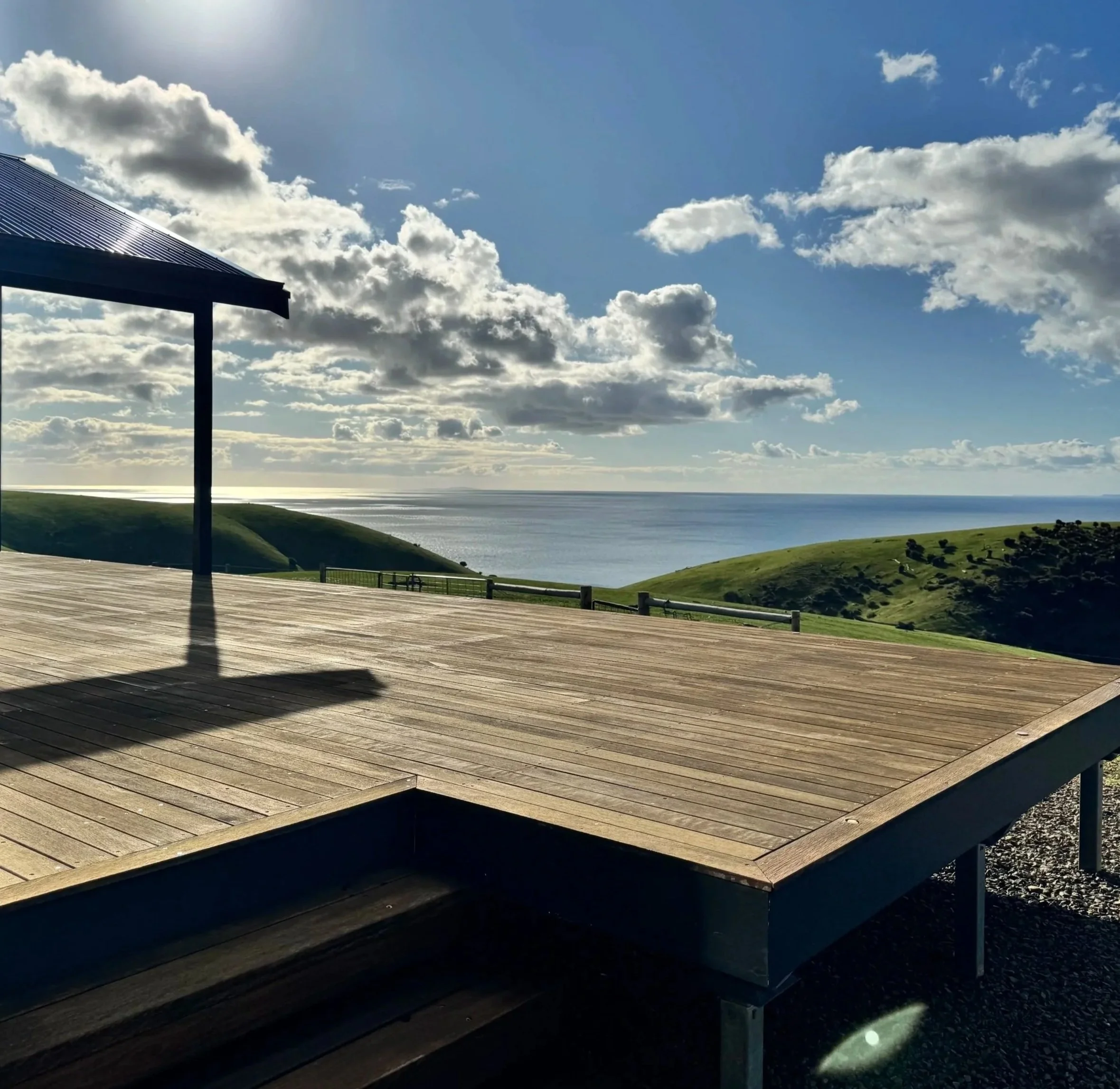 A wooden deck overlooking green hills and the ocean under a partly cloudy sky.