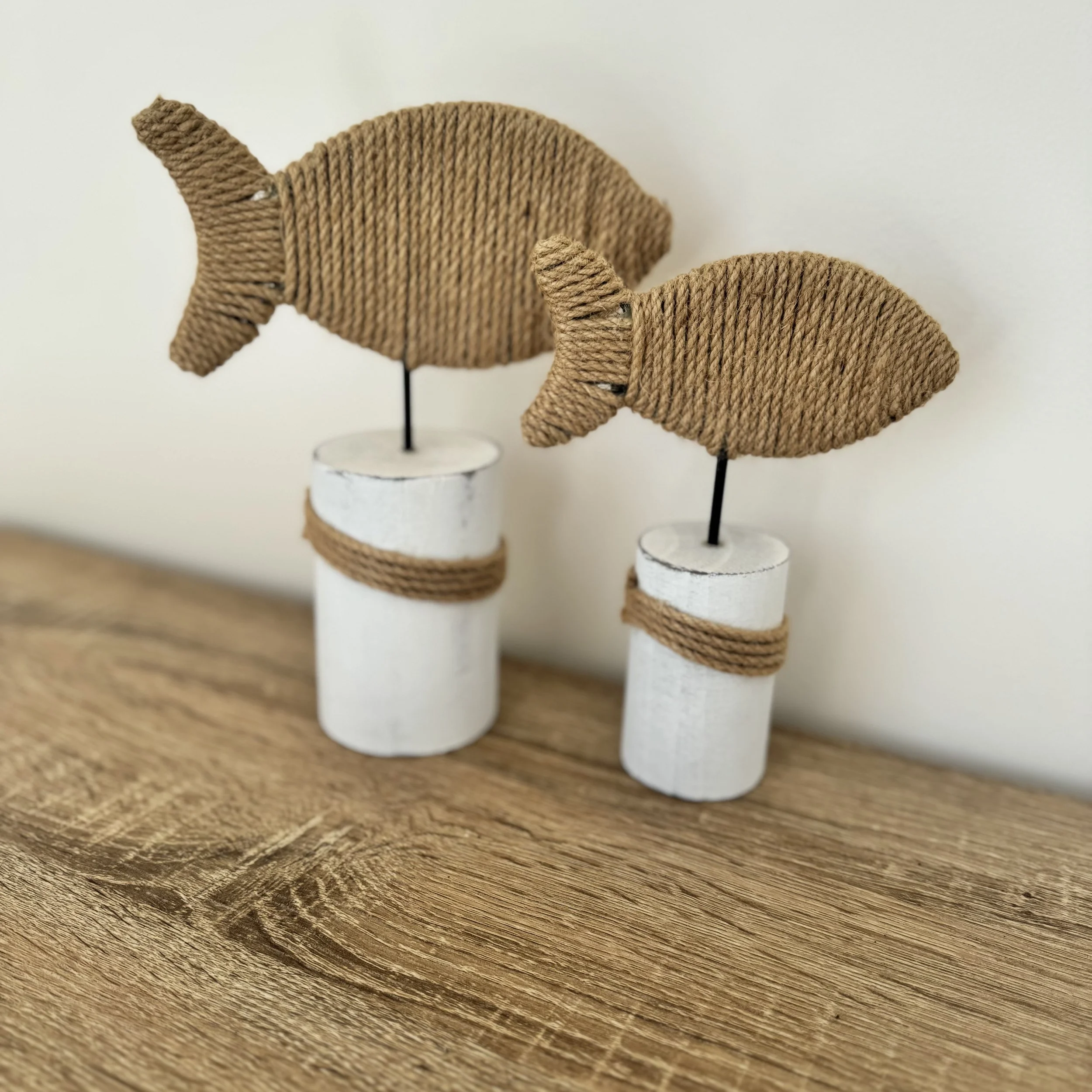 Decorative fish-shaped figures made of woven material, mounted on black rods and standing on white cylindrical bases with rope accents, placed on a wooden surface.