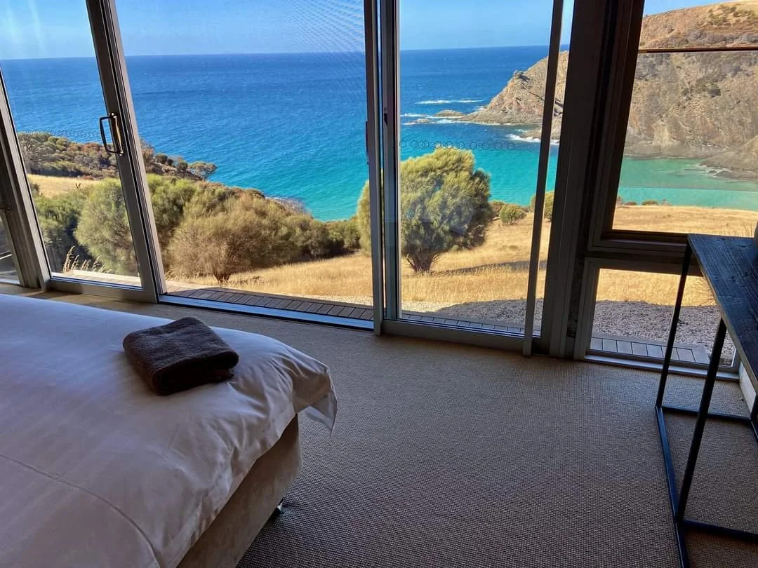 A bedroom with a large window overlooking a coastal landscape with blue ocean, green trees, and cliffs.