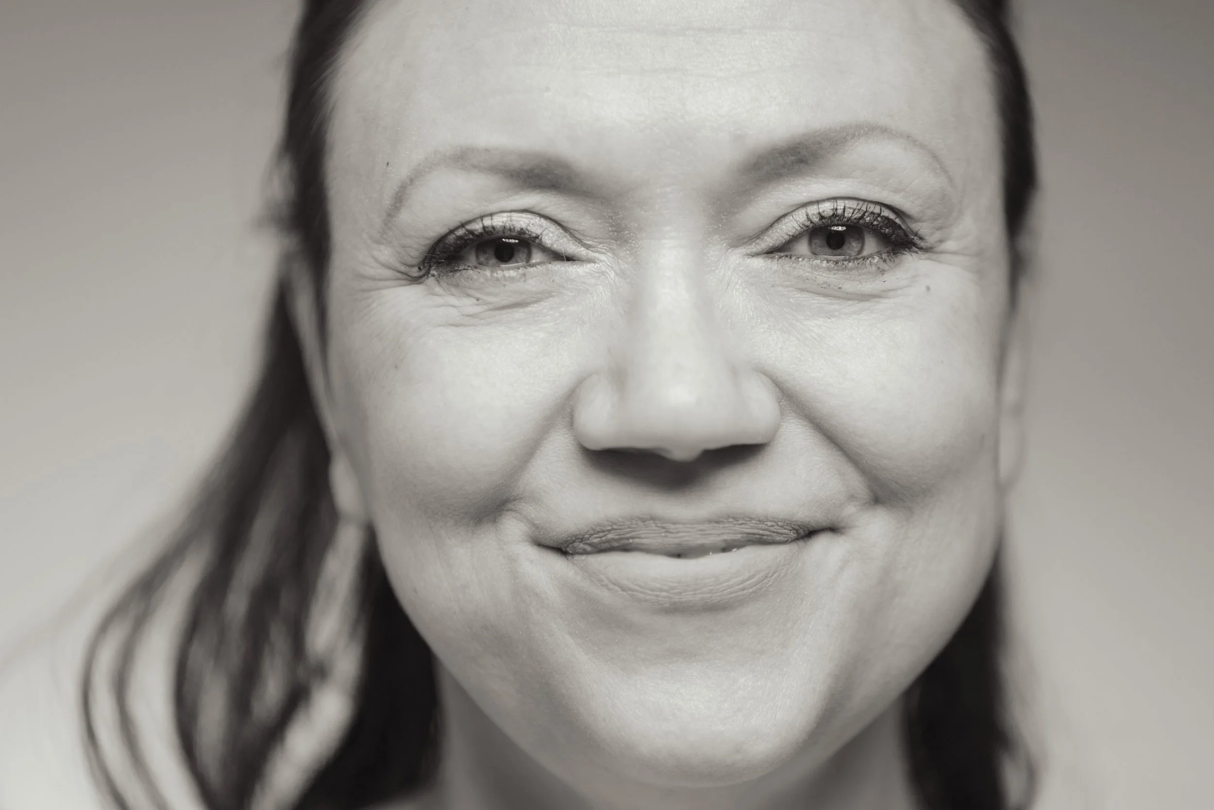 A close-up black and white photograph of a woman's face with a subtle smile, showing her eyes, nose, and mouth, with smooth skin and dark hair.