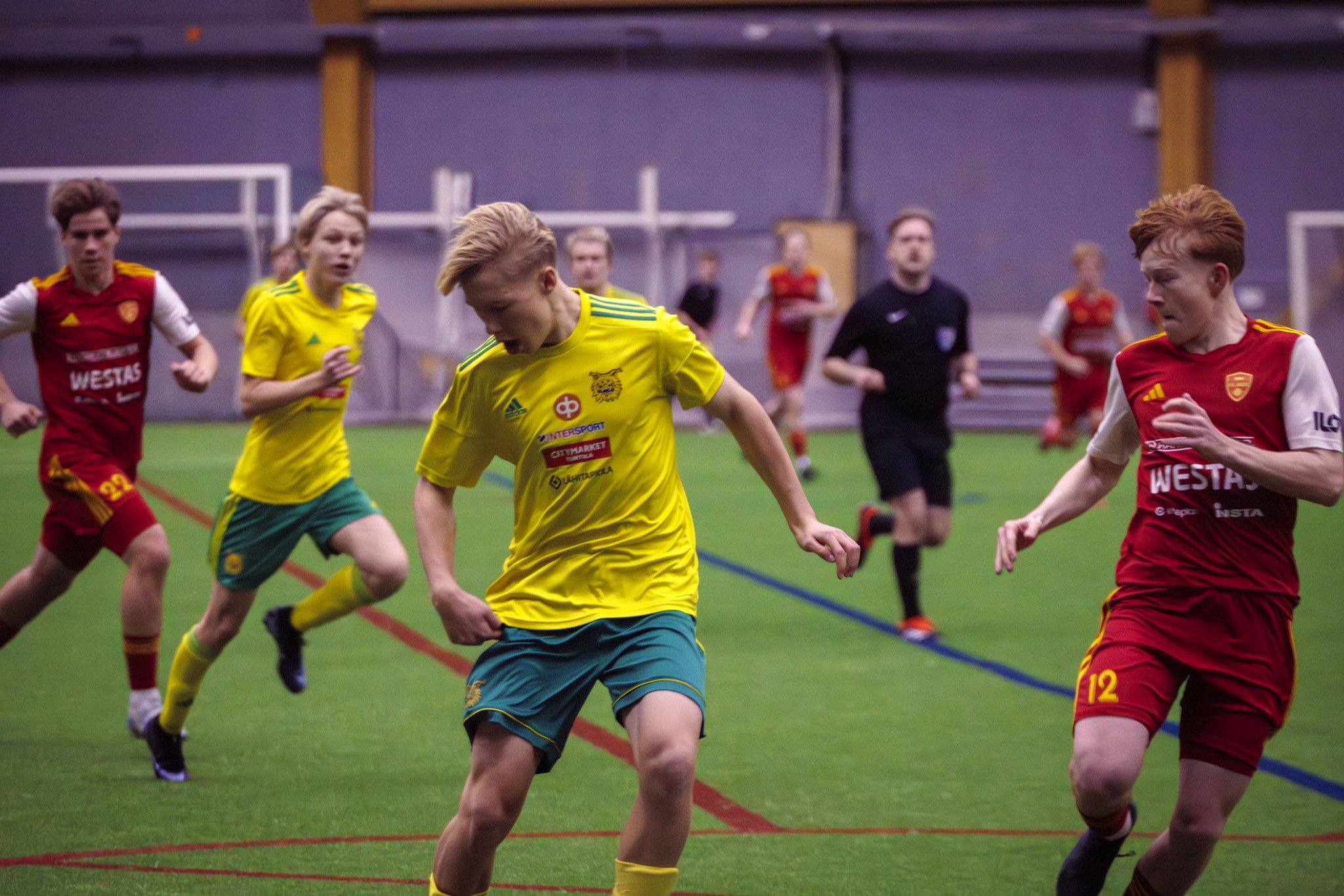 Young male soccer players in a match on an indoor field, with players in yellow and red jerseys running and chasing the ball.